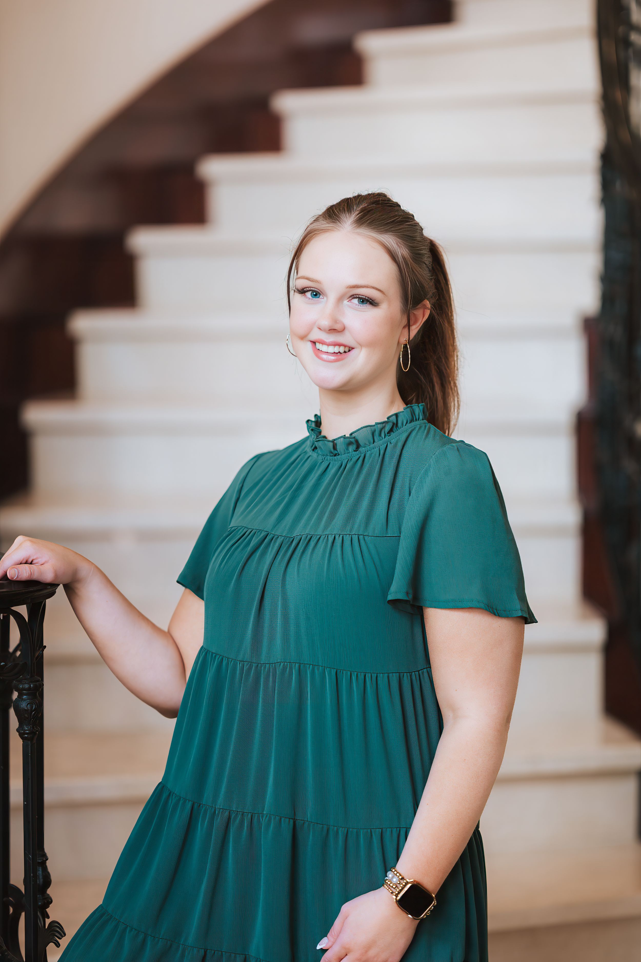 Young woman in green tiered dress smiling on elegant staircase, wearing gold watch and hoop earrings.