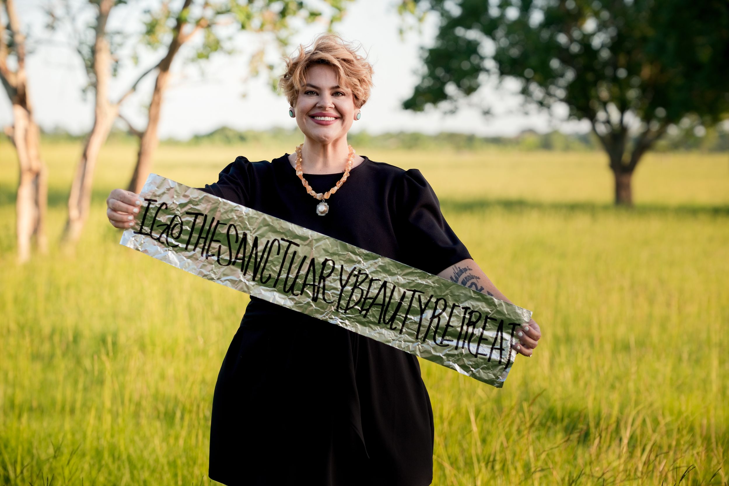 Woman in black dress holding a decorative sign in a lush green field with trees in the background.