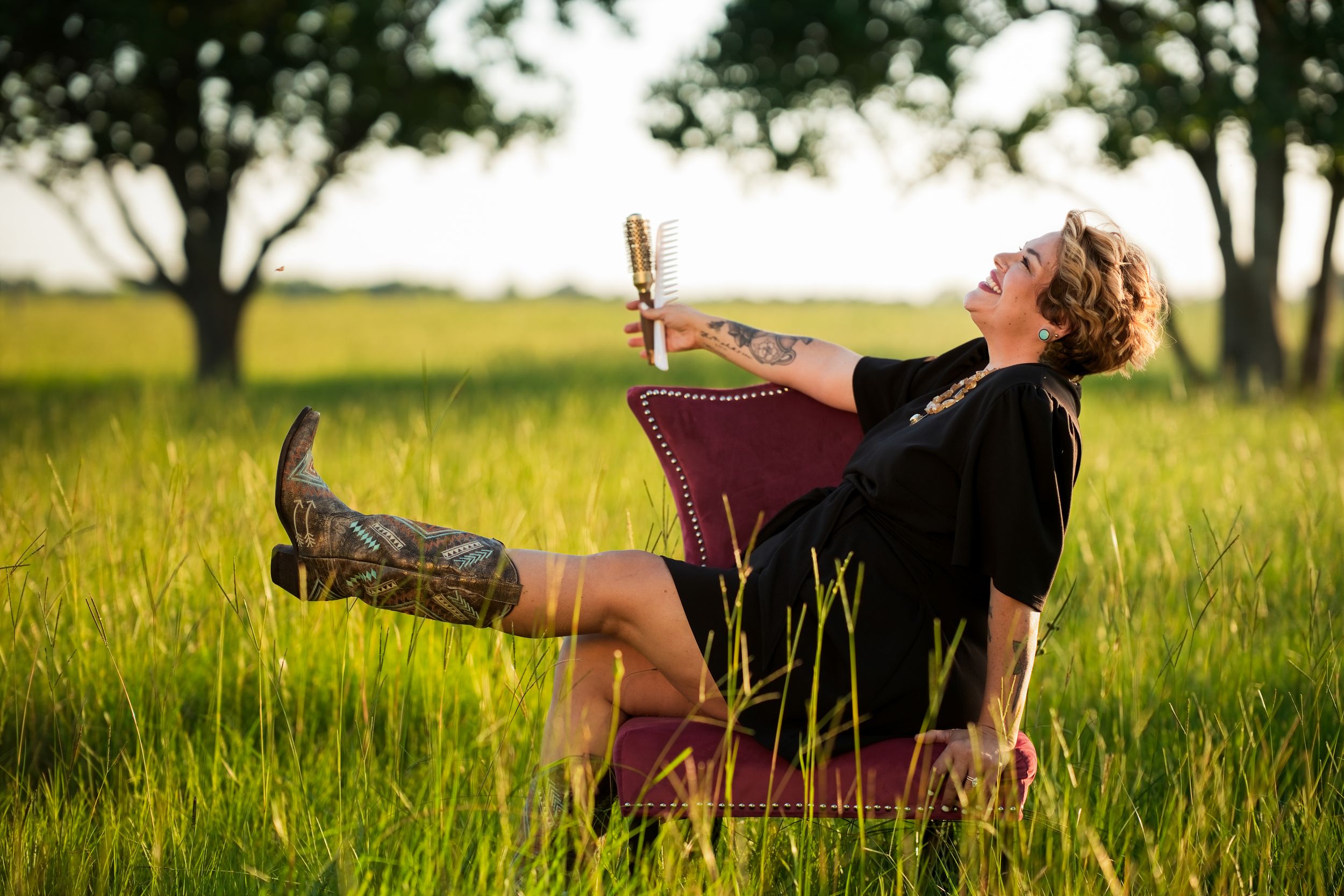 Woman with tattoos relaxing in a red chair in a green field, wearing cowboy boots and holding a microphone.