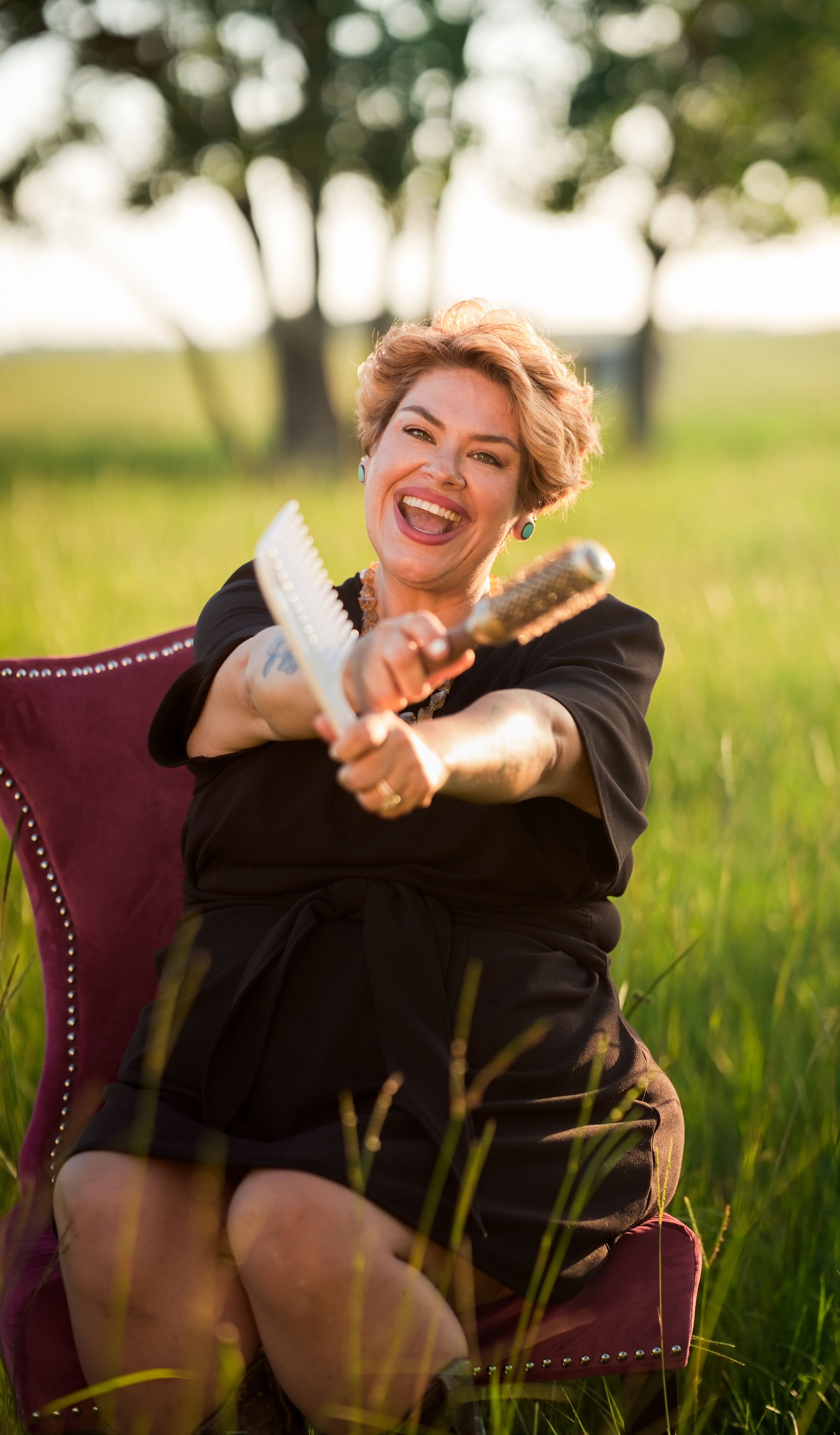 Smiling woman in black dress sitting in red chair outdoors, holding award trophy in sunny green field.