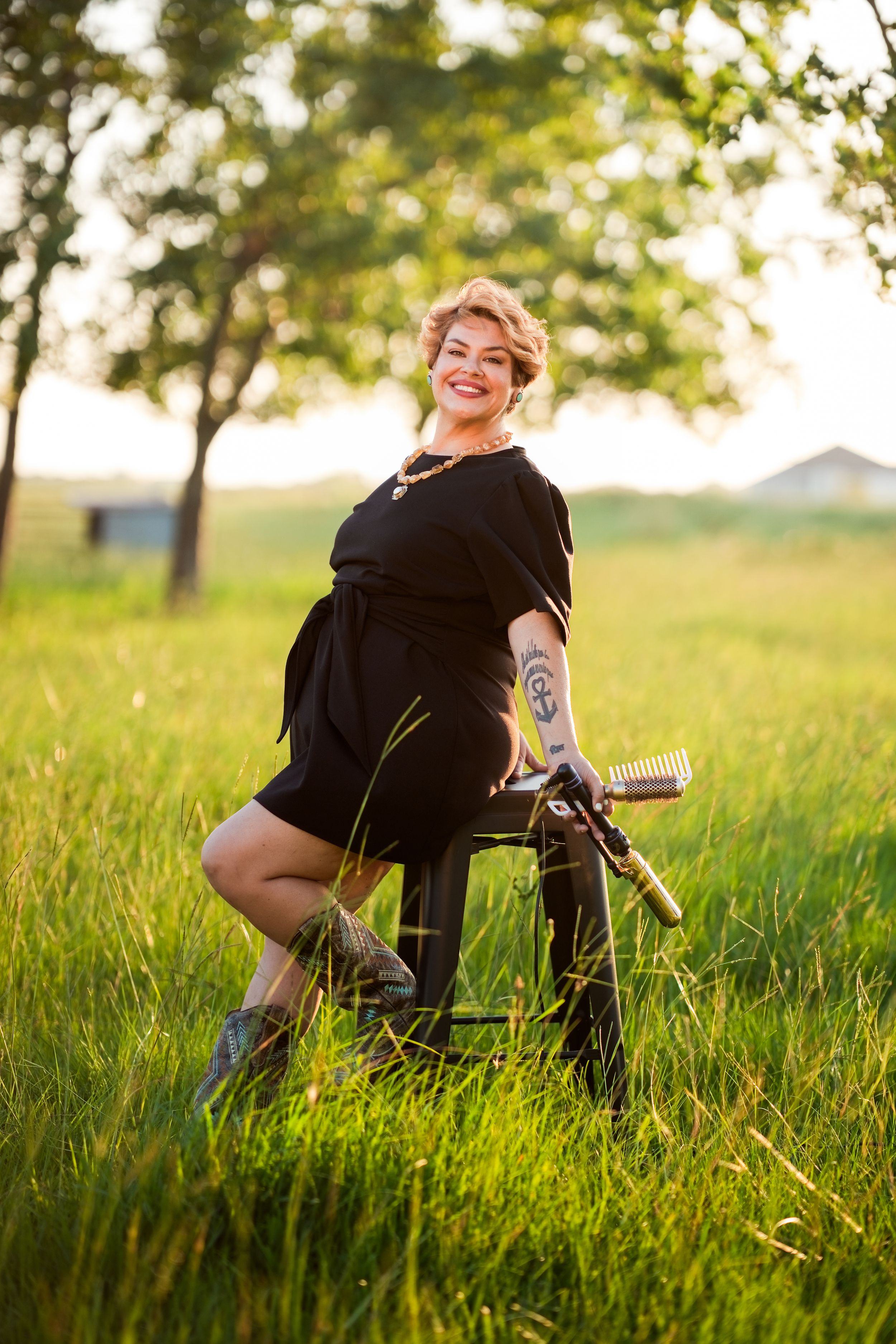 Smiling woman with tattoos in black dress sits on stool in sunny green field with trees in background.