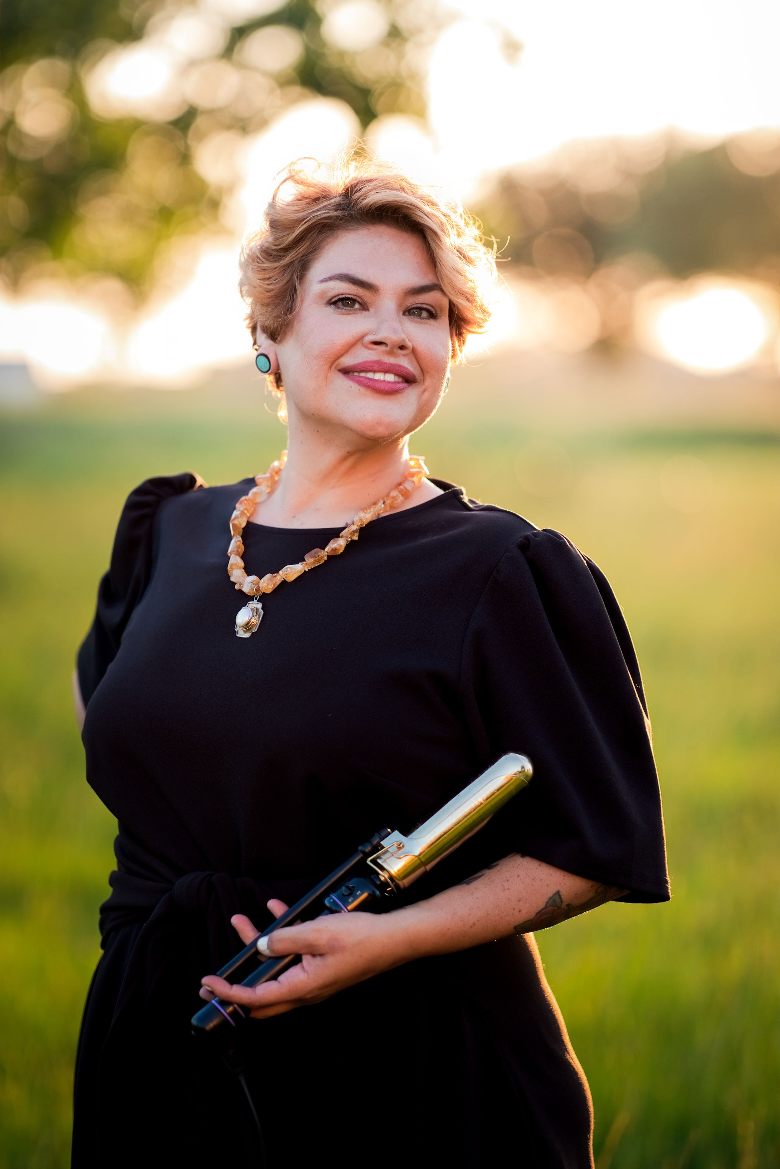 Woman in black dress holding wine bottle outdoors at golden hour, wearing amber necklace and turquoise earrings.