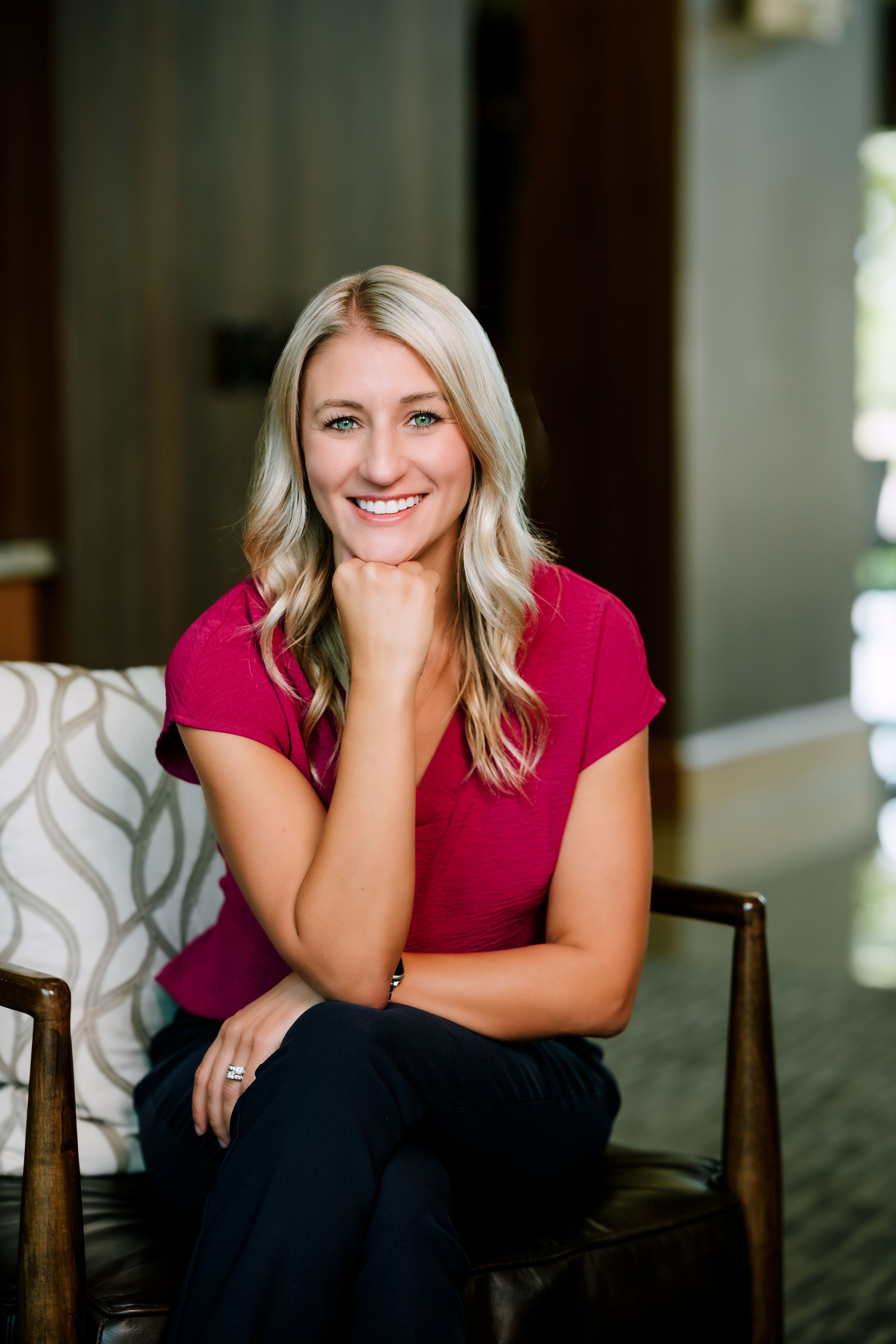 Smiling blonde woman in a magenta top sitting confidently in a chair with hand on chin in a modern interior setting.