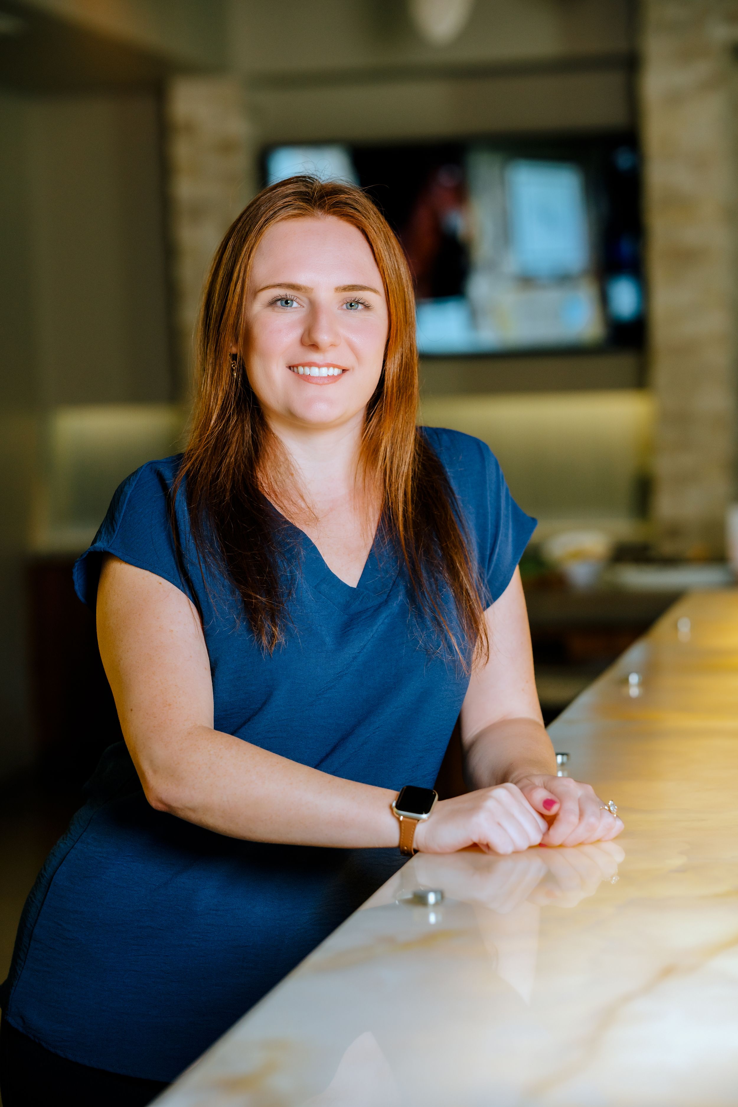 Smiling woman with red hair in blue scrubs leaning on a marble counter in a modern medical office.