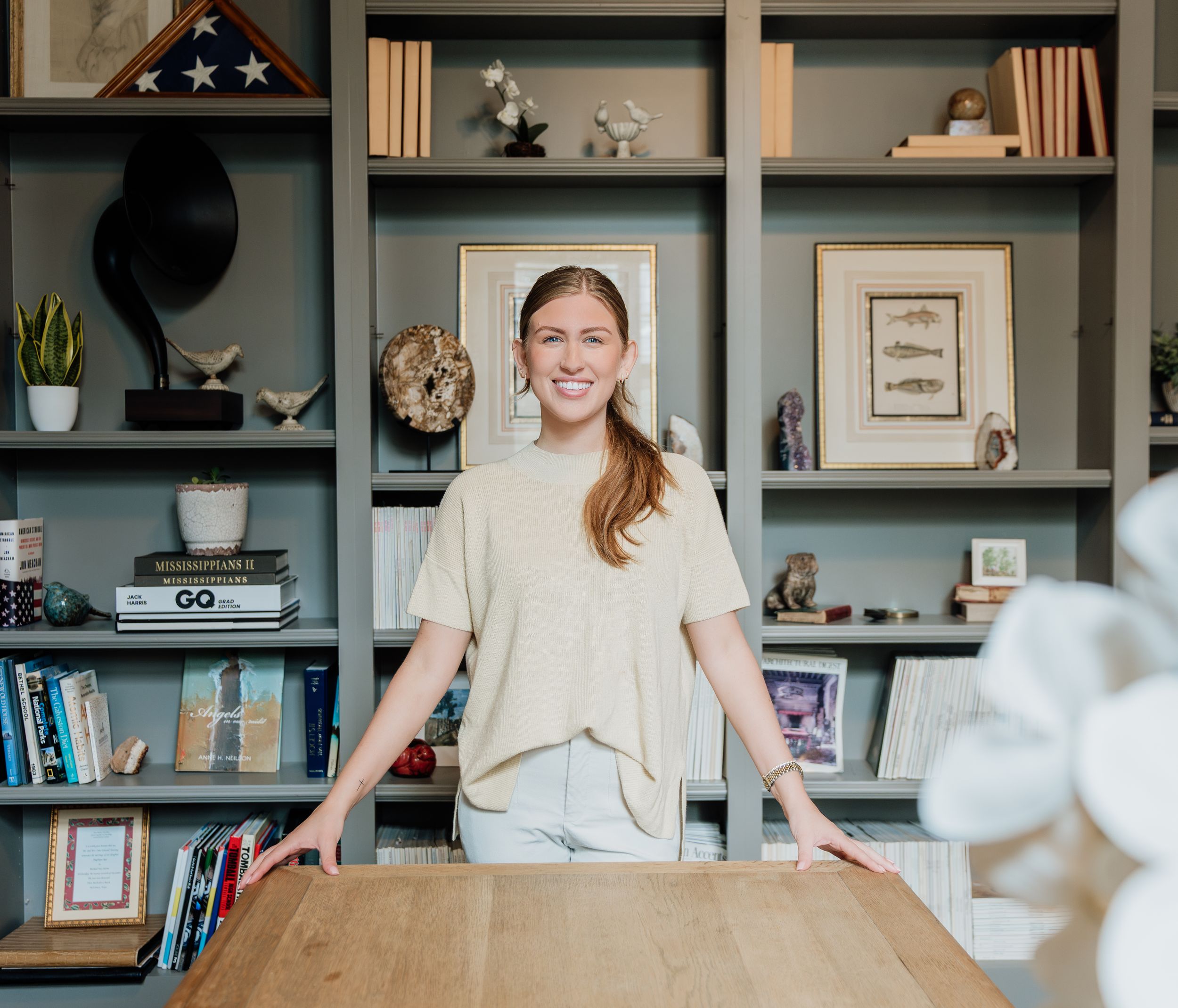 Smiling woman standing behind a wooden desk in front of styled bookshelves with books and decor.
