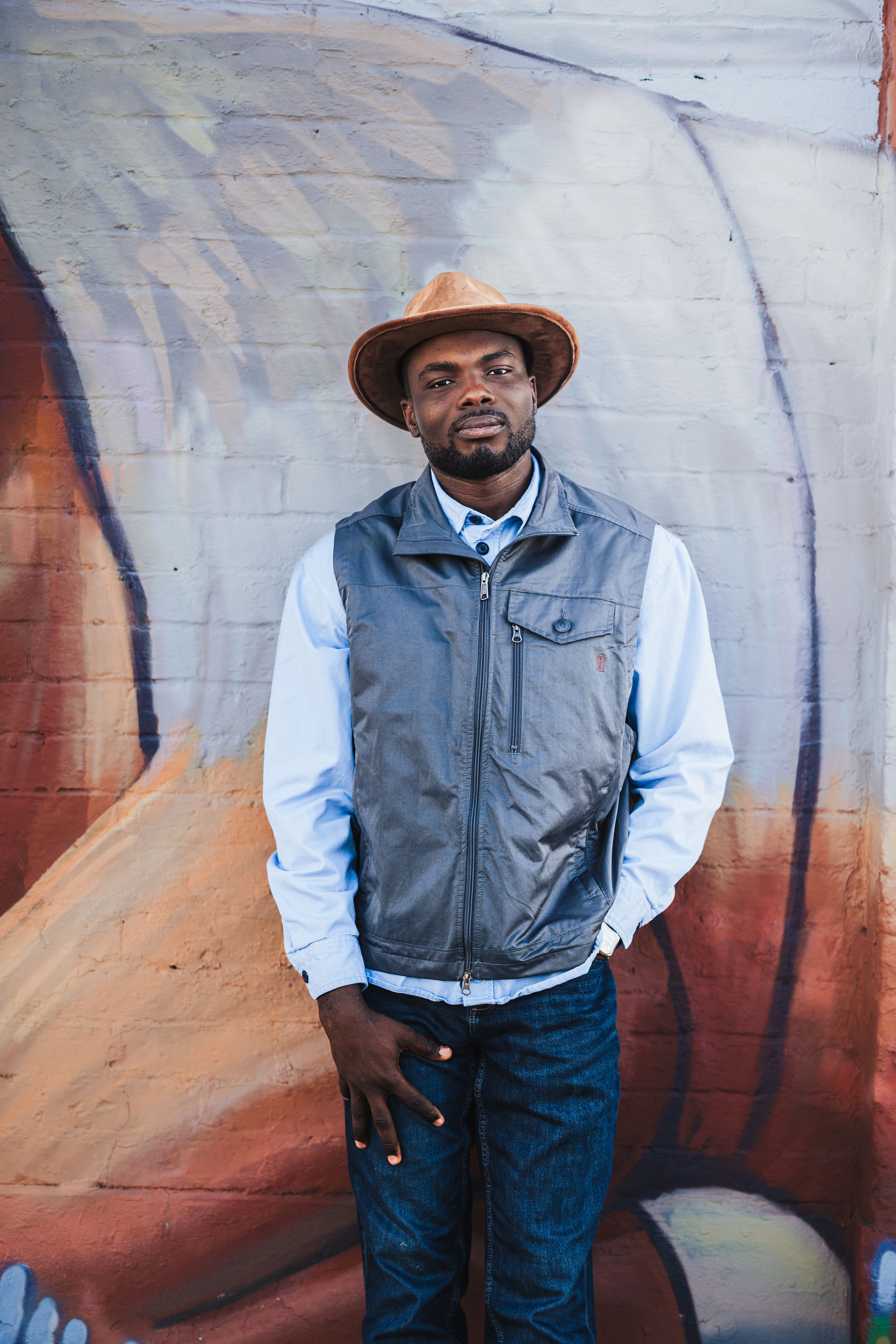 Man in brown hat, gray vest, and blue shirt stands confidently before a colorful mural wall.