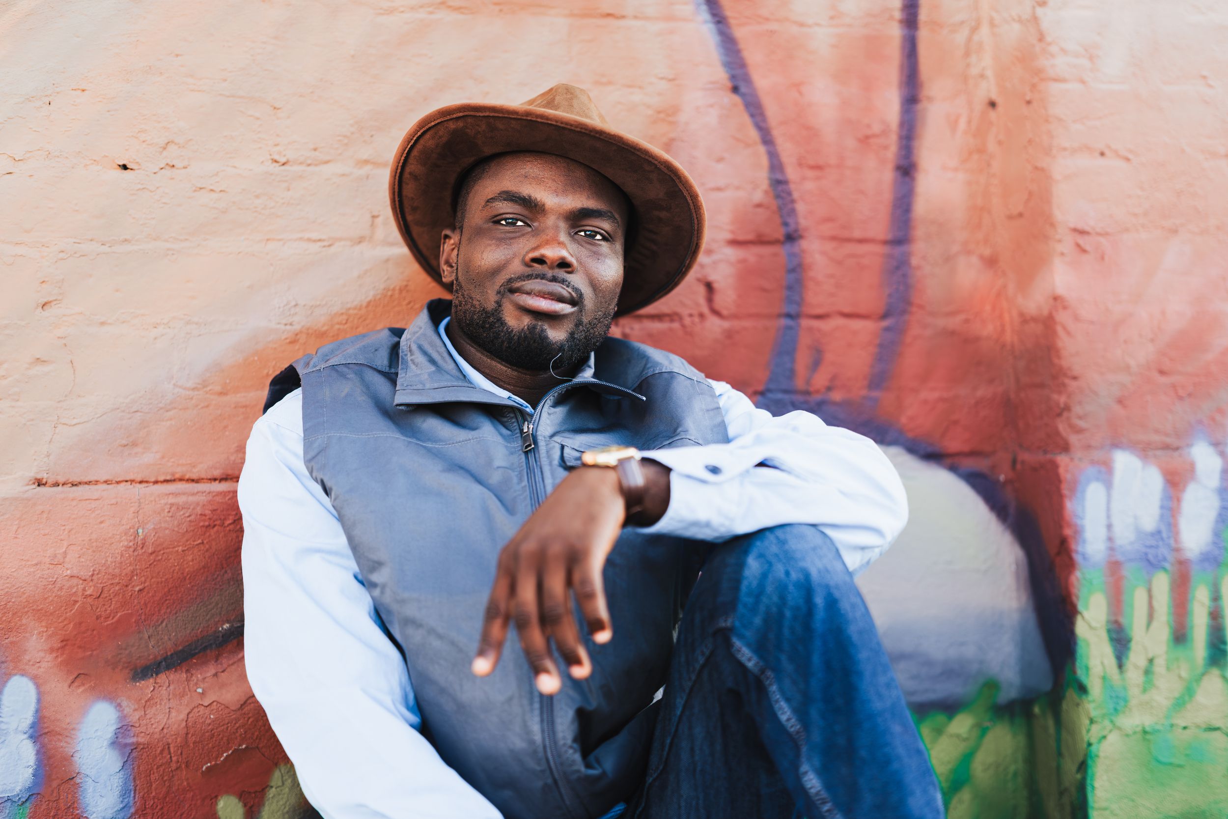 Man in brown hat and vest sits relaxed against colorful graffiti wall outdoors.