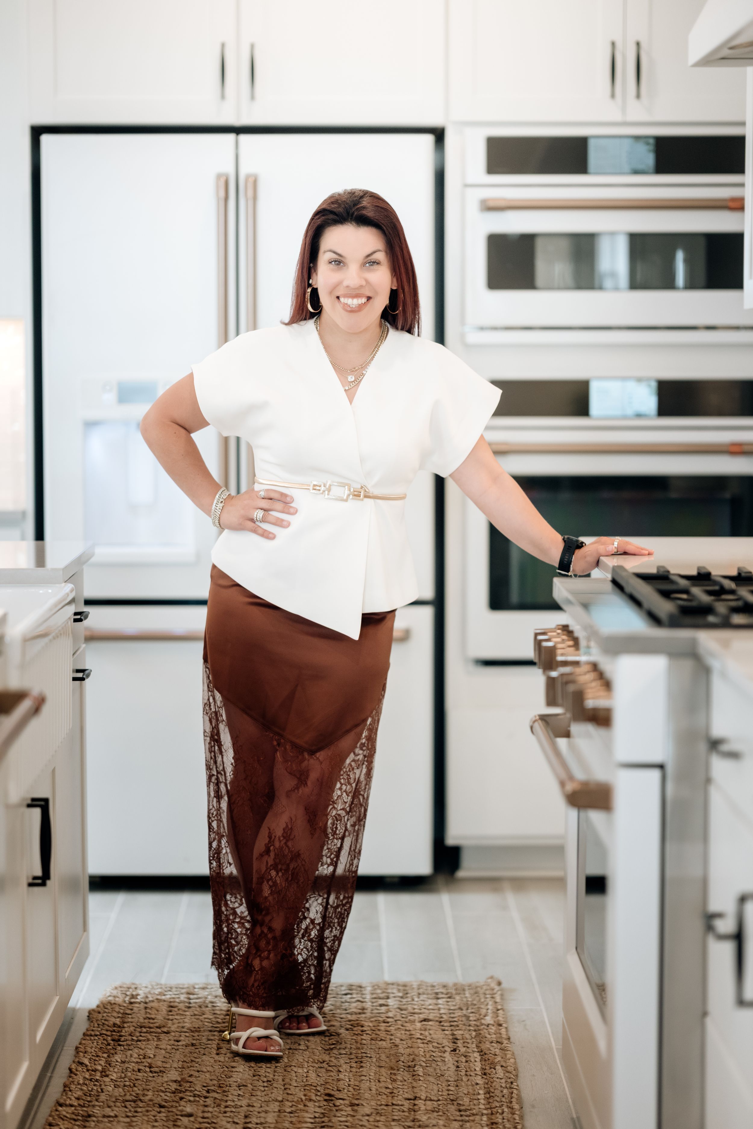Woman in white top and brown skirt posing confidently in a modern white kitchen with stainless steel appliances.