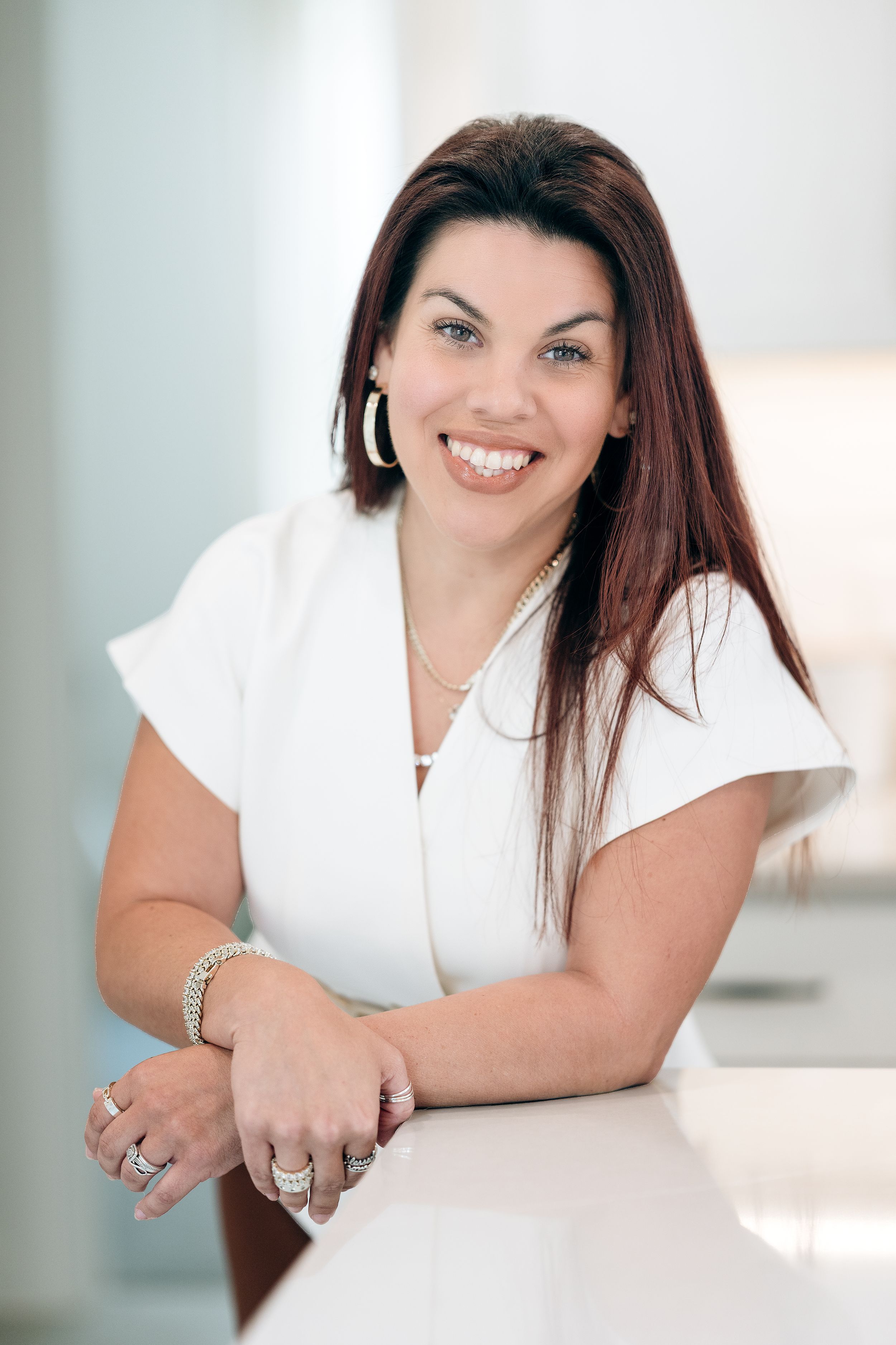 Smiling woman with long dark hair wearing a white dress and jewelry, leaning on a white surface in a bright modern setting.