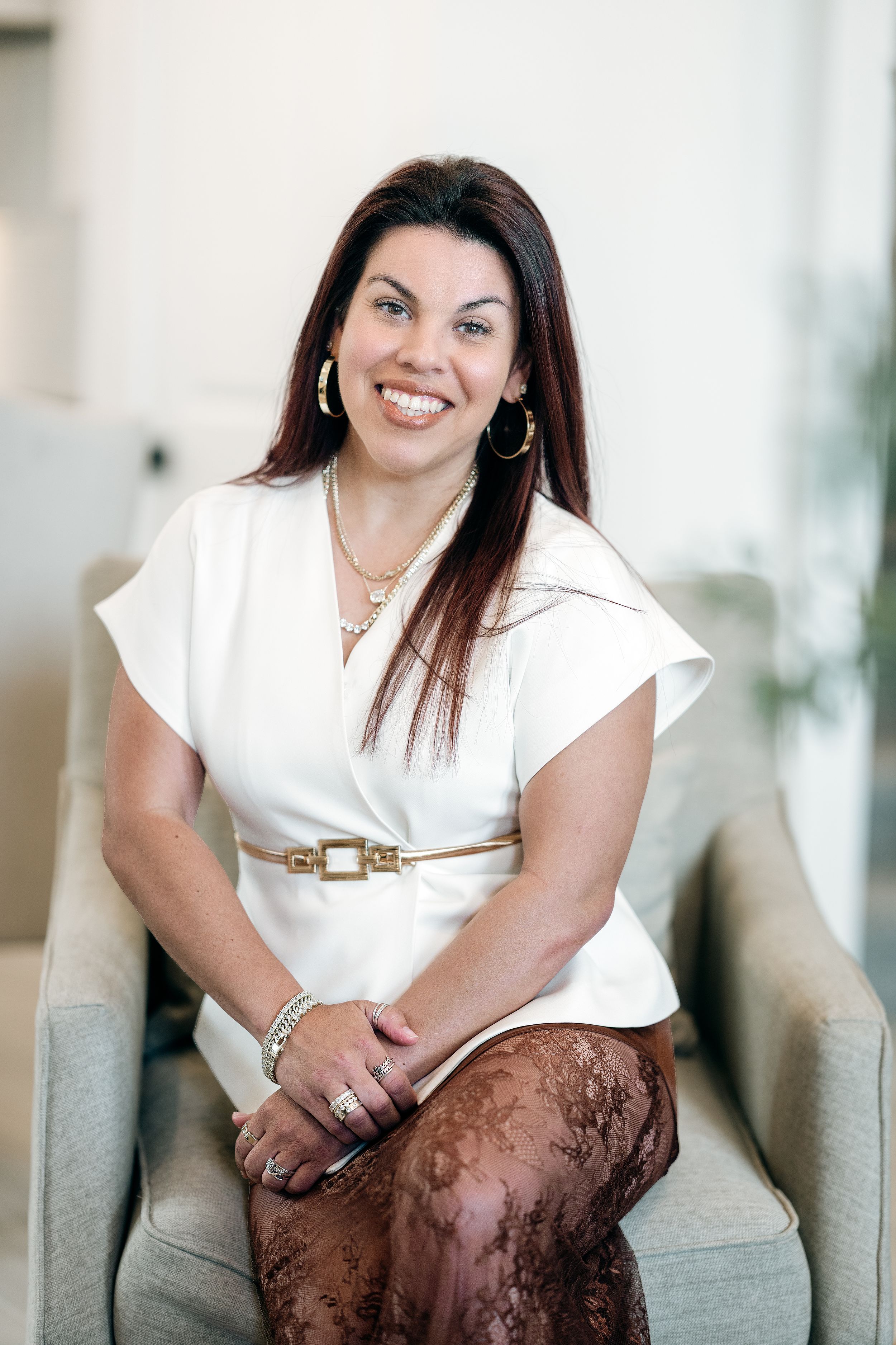 Smiling woman in white top with gold belt sits in chair, wearing hoop earrings and layered necklaces.
