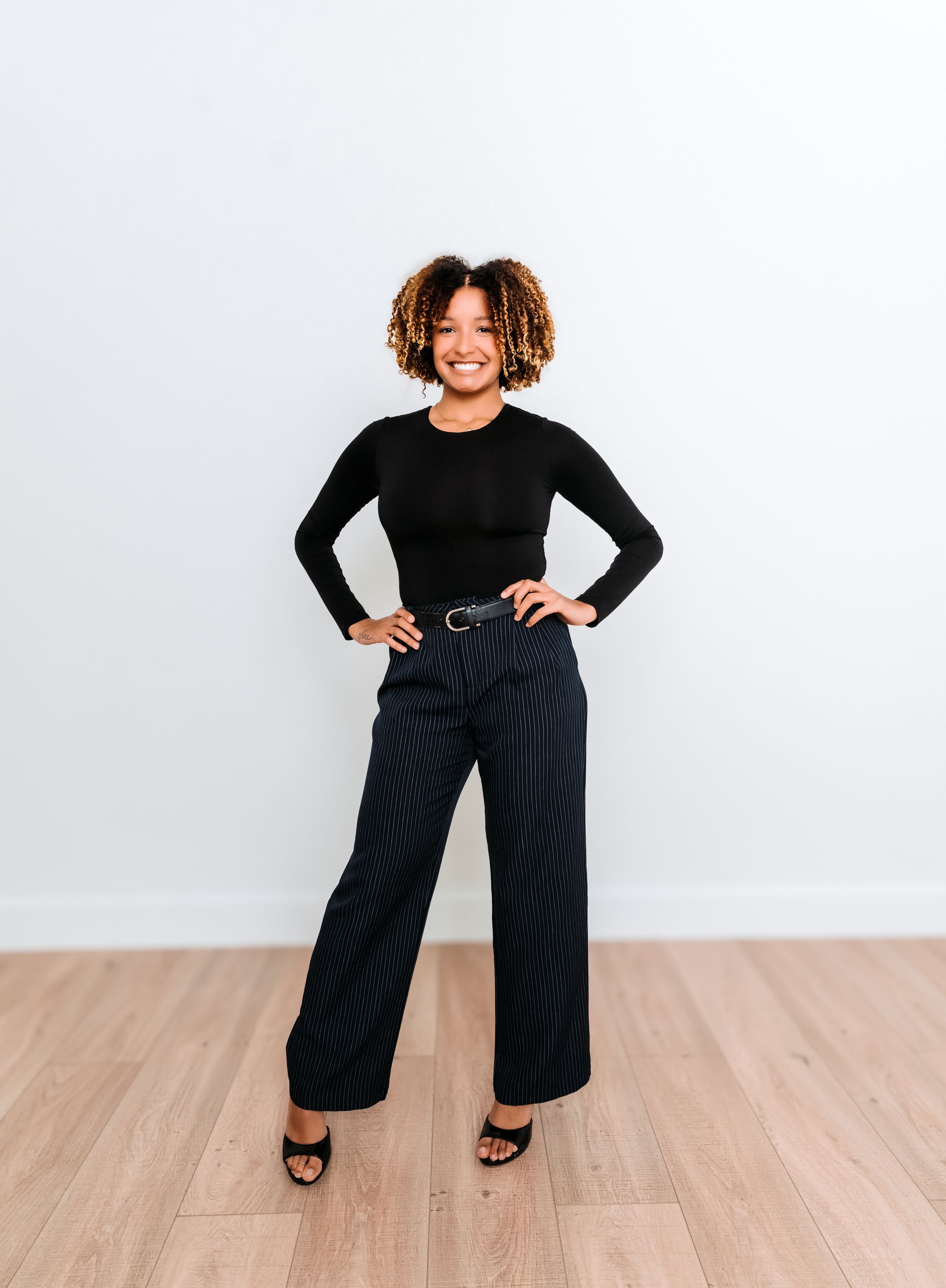 A person in an all-black outfit strikes four different poses on a light wooden floor against a white wall.