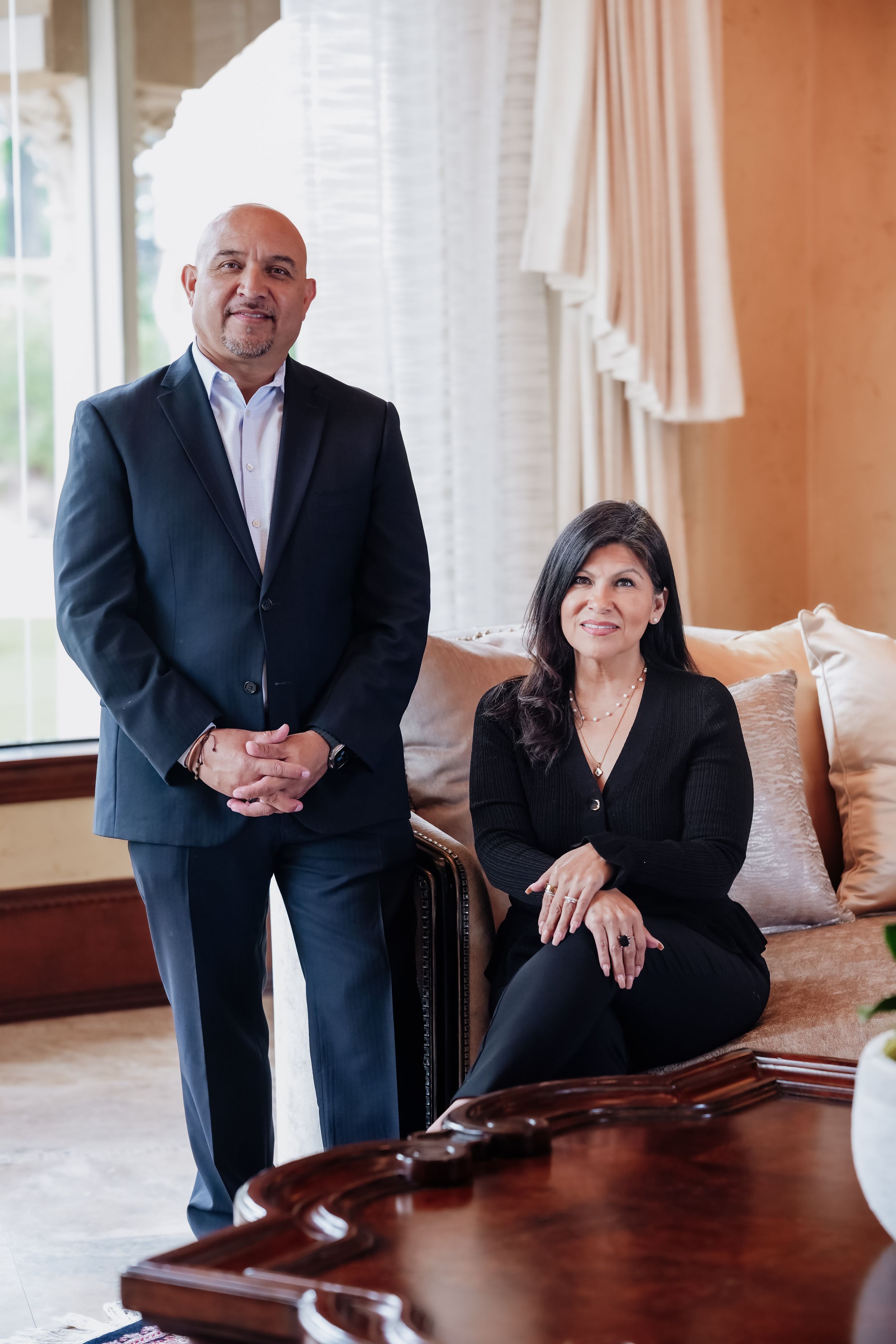 A man in a dark suit and a woman in black attire pose professionally in an elegant living room setting.