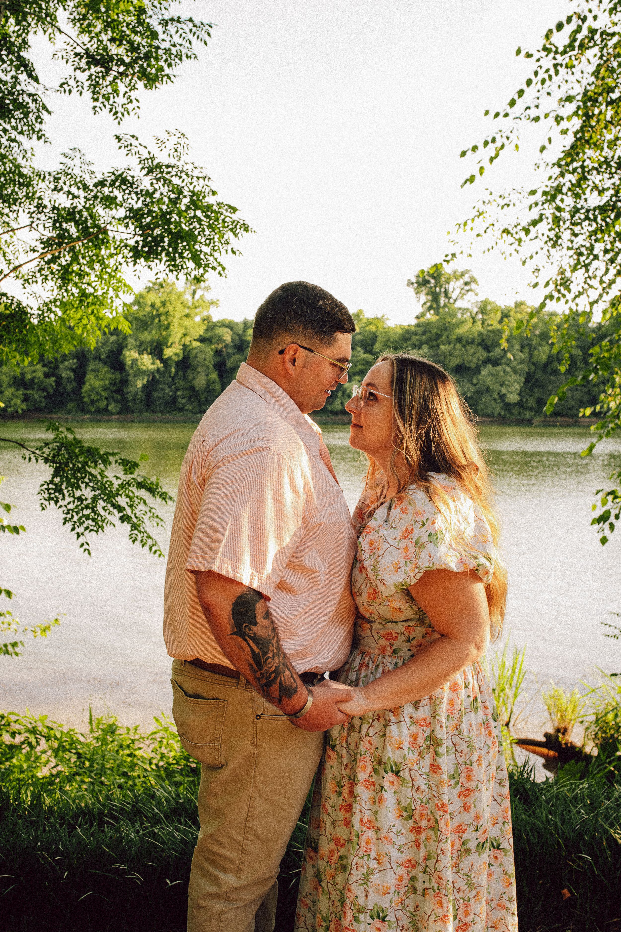An intimate couple embraces by a serene lake with overhanging tree branches in golden light.