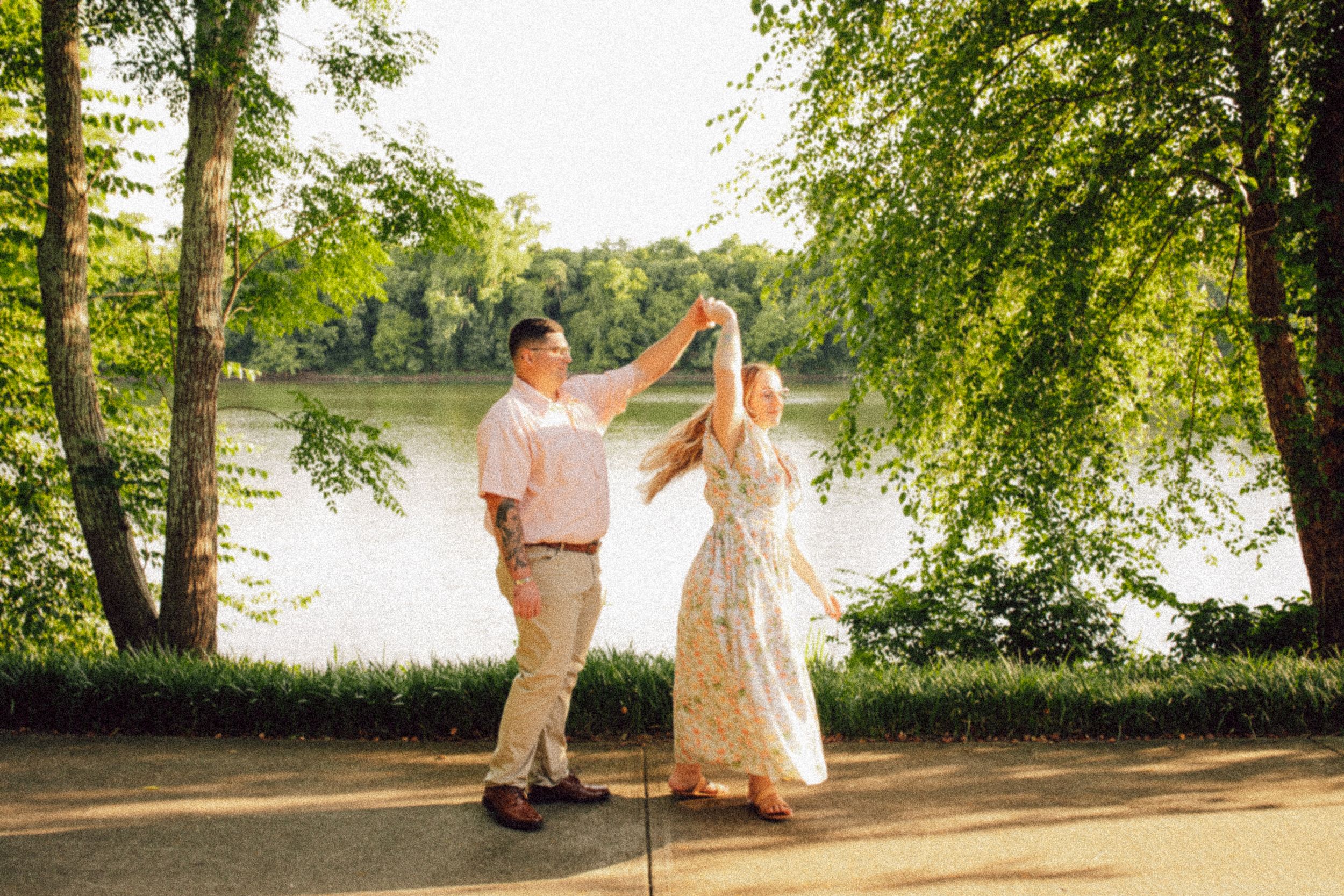 A romantic dance pose by a peaceful lake surrounded by lush green trees at sunset.