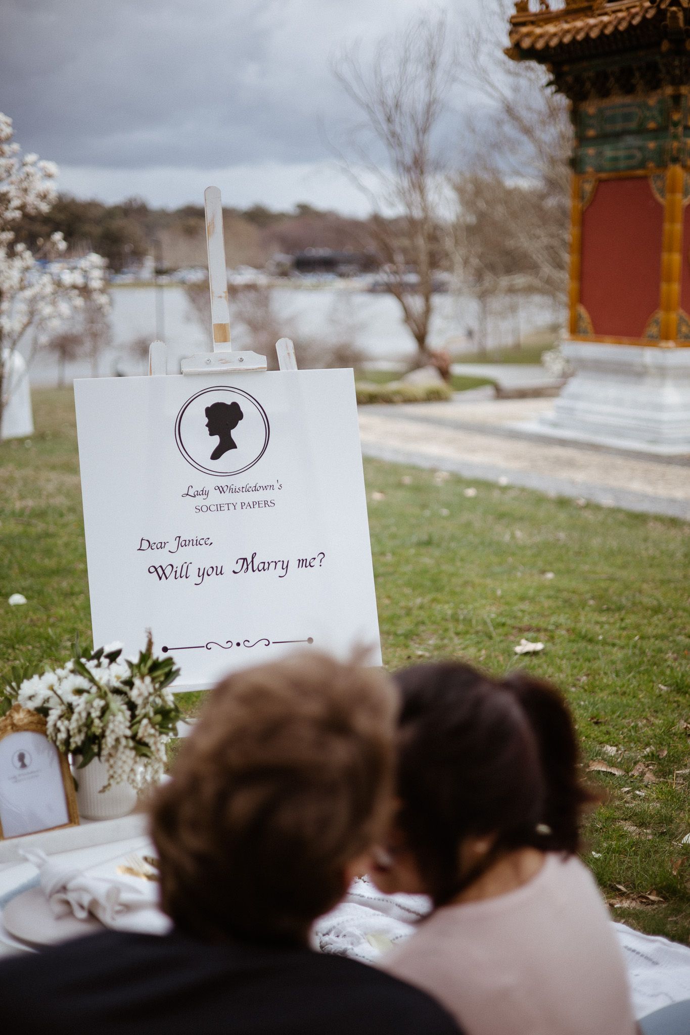 A marriage proposal setup outdoors with a silhouetted couple viewing a handwritten note and decorated picnic near a pagoda.