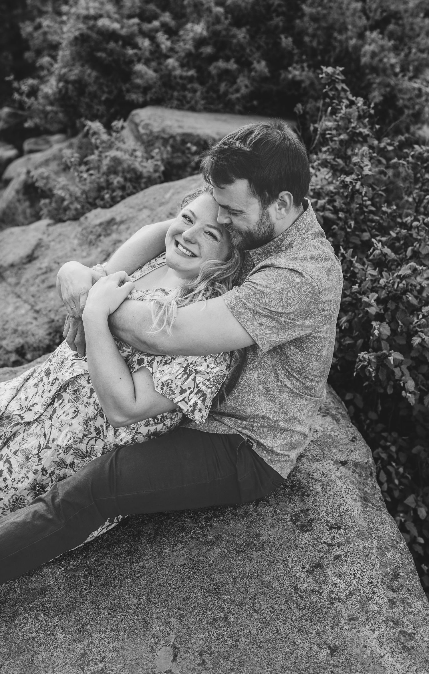 A romantic black and white photo of a couple embracing and laughing on a rock formation.