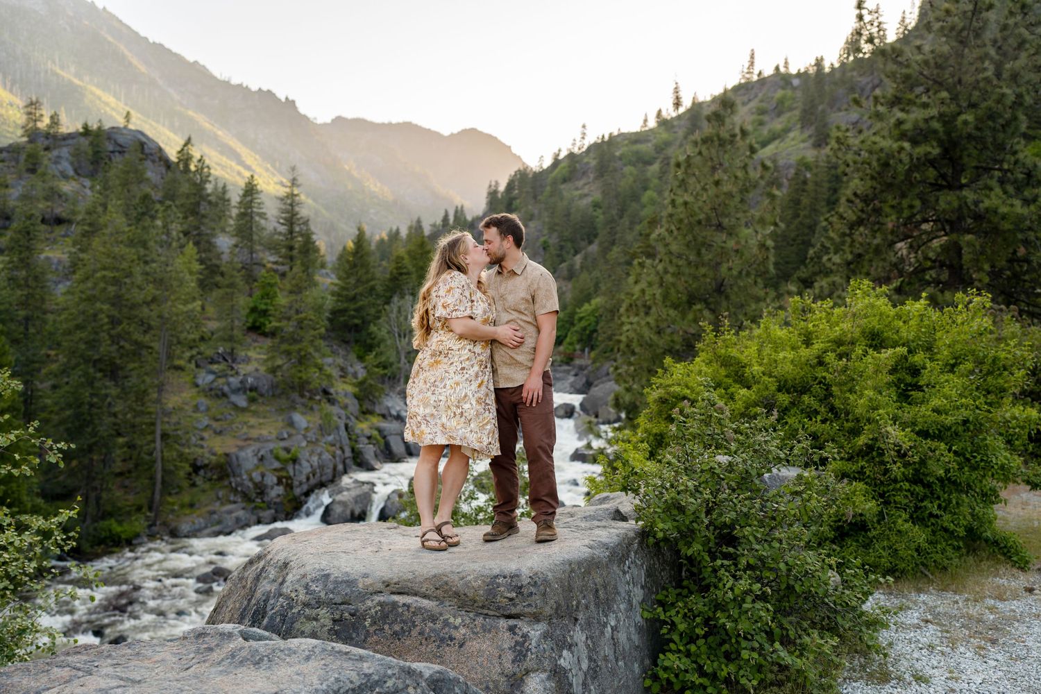 A couple stands on a large rock overlooking a mountain stream in a scenic wilderness setting.
