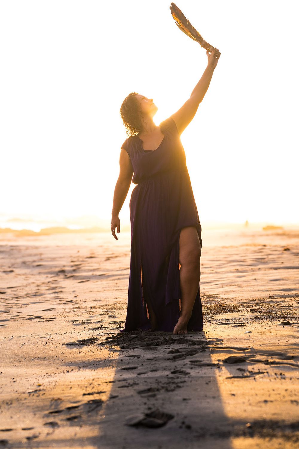 Figure in flowing dress poses dramatically on beach during golden hour sunset.