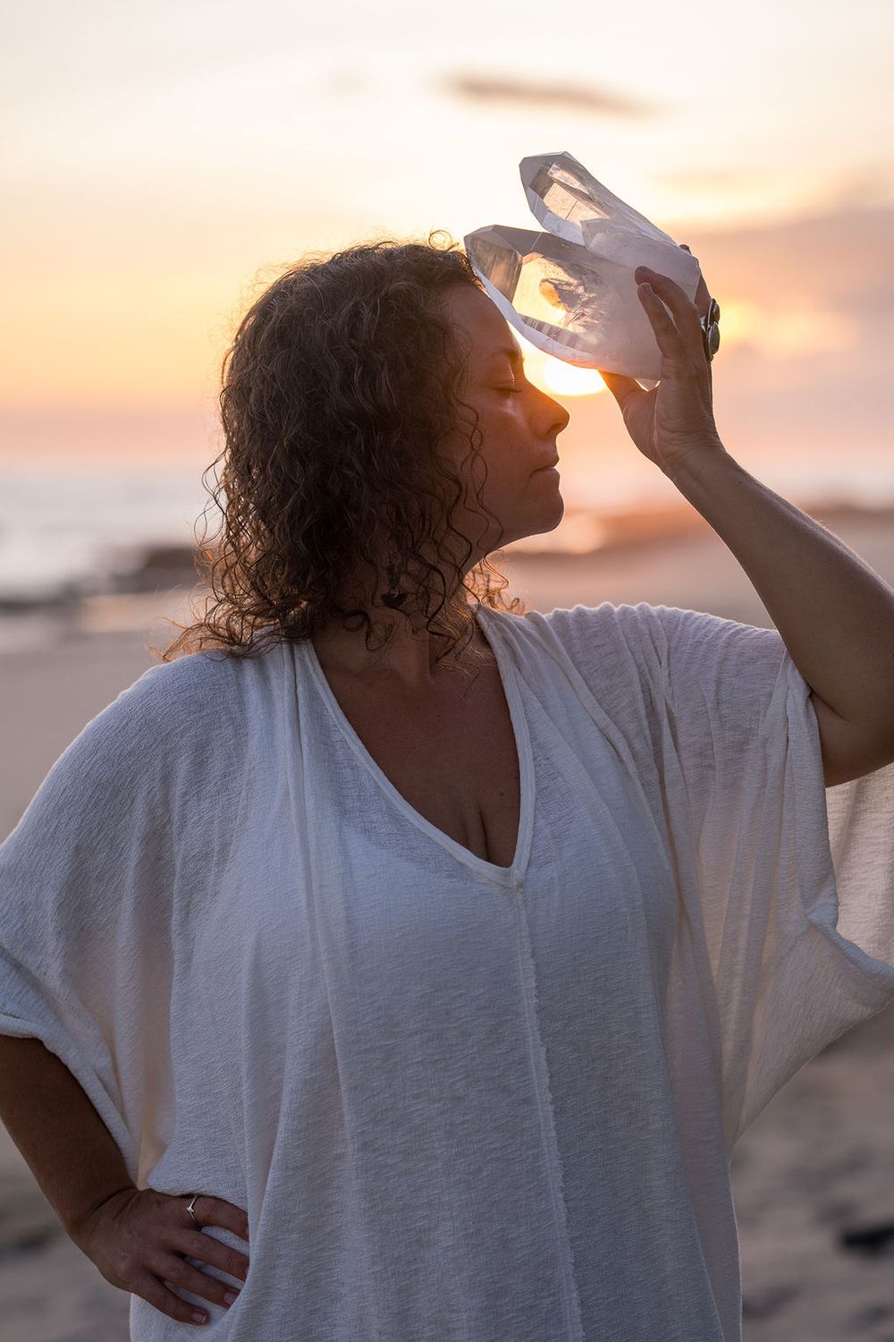 Person in white top holds crystal against sunset sky at beach.