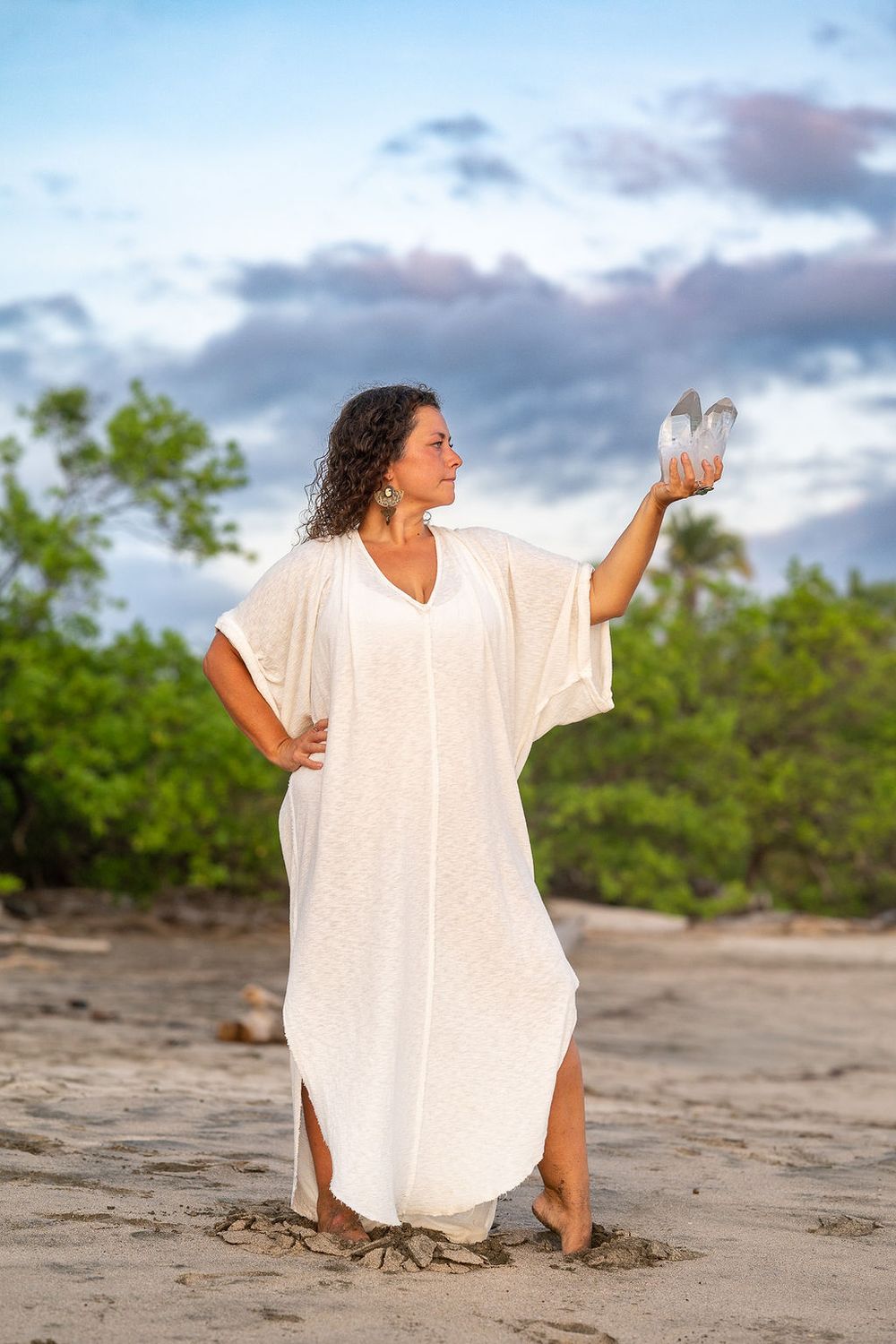 Figure in flowing white dress holds crystal while standing on sandy beach.