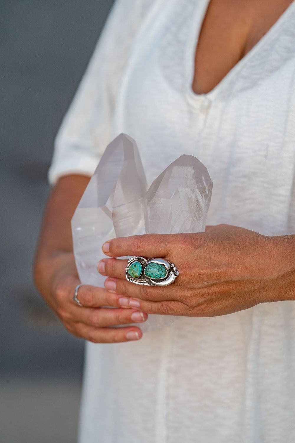 Close-up of hands holding clear quartz crystals with a turquoise statement ring against a white flowy outfit.