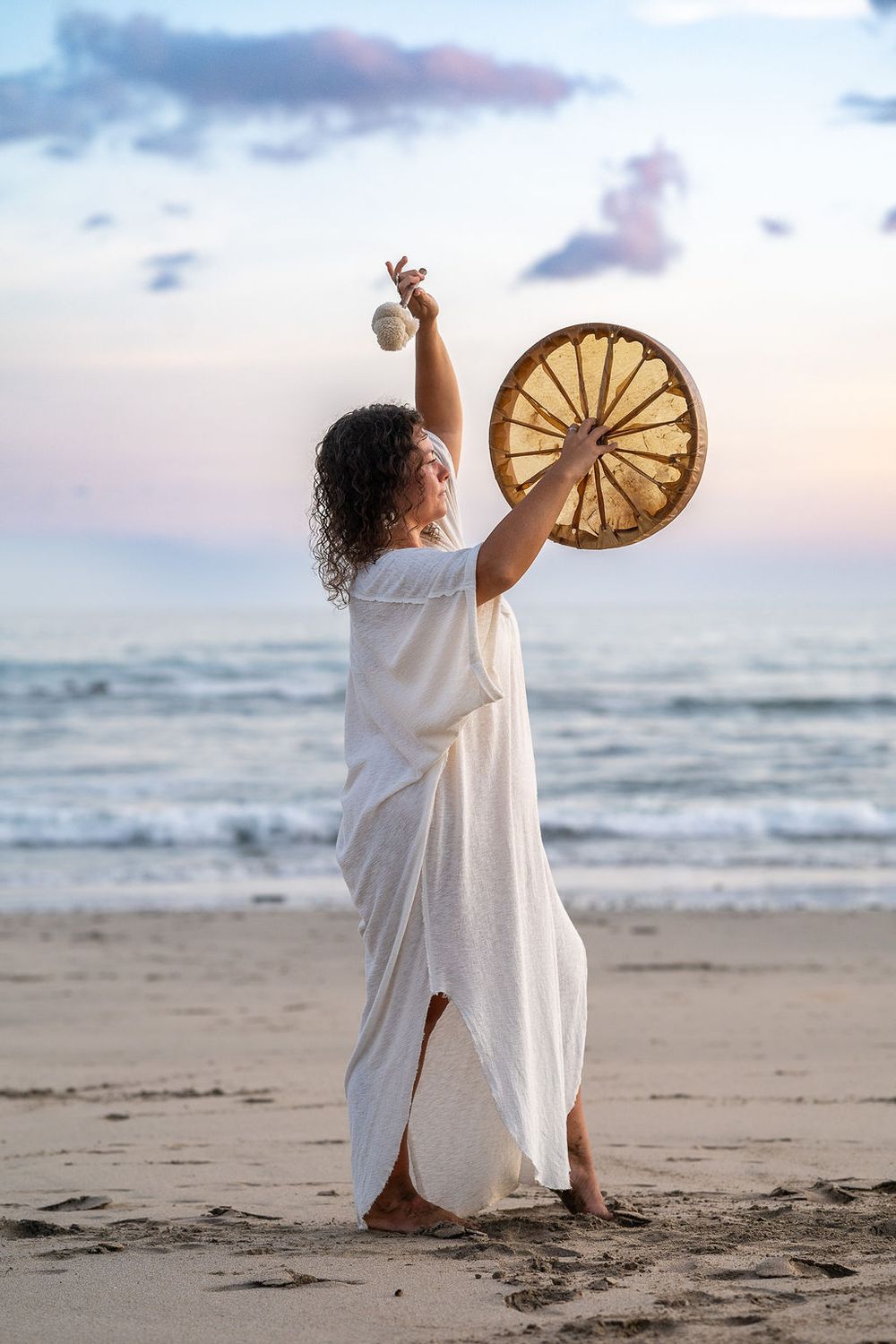 Figure in flowing white dress holding a ceremonial drum and sage bundle on beach at sunset.