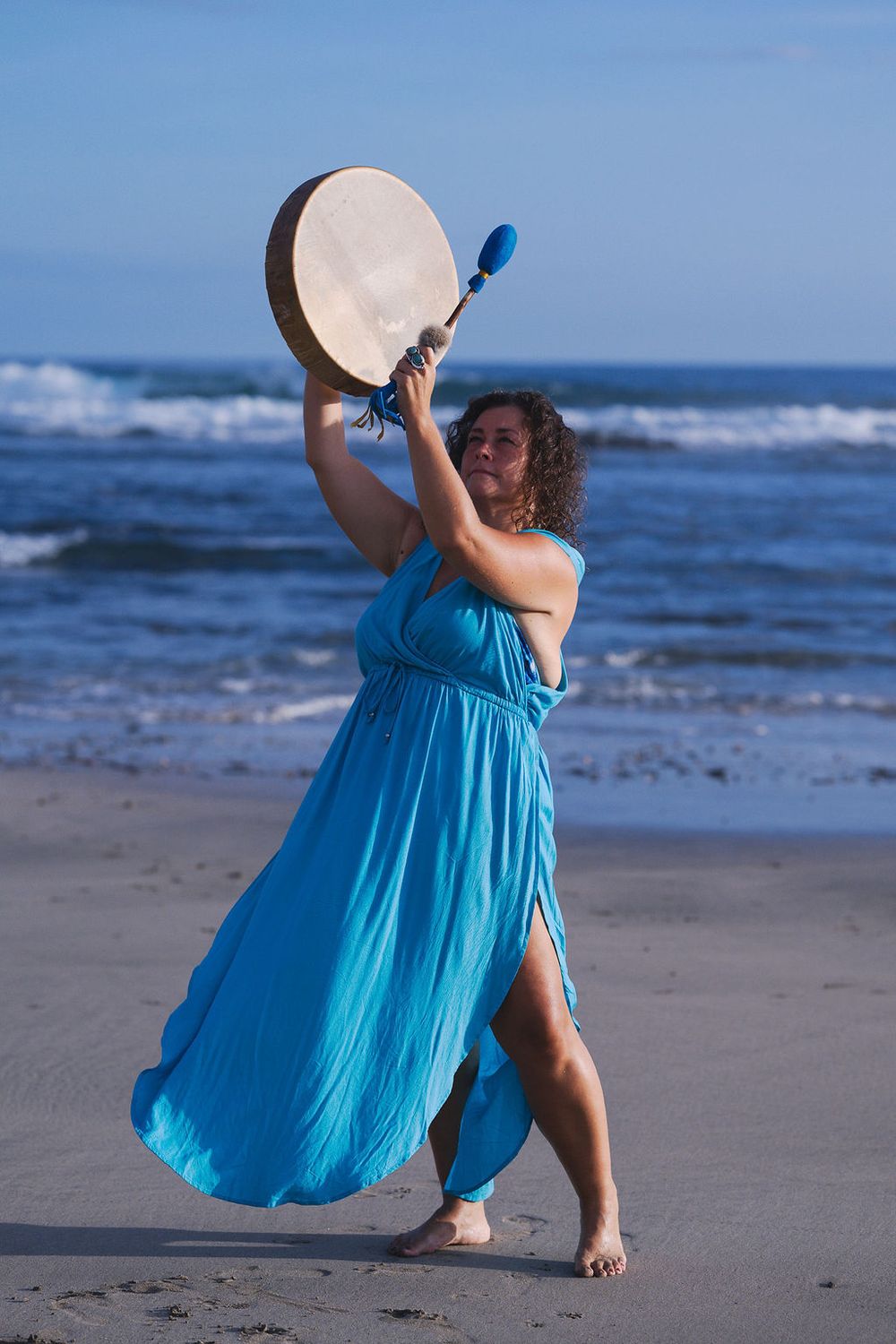 Dancing on beach with tambourine in flowing turquoise dress against ocean waves.
