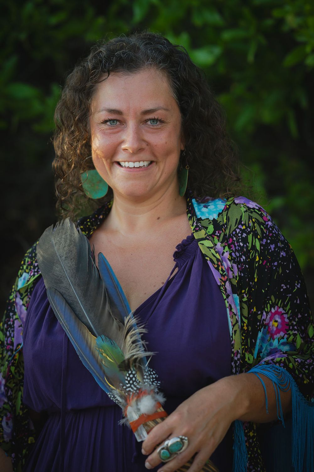 A person in a purple floral dress holds feathers while posing outdoors against green foliage.