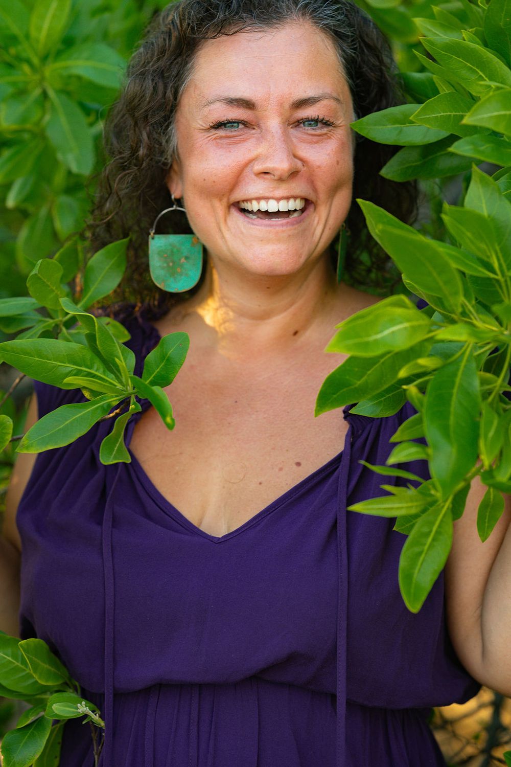 Someone wearing turquoise earrings and purple top smiles brightly among green leaves.