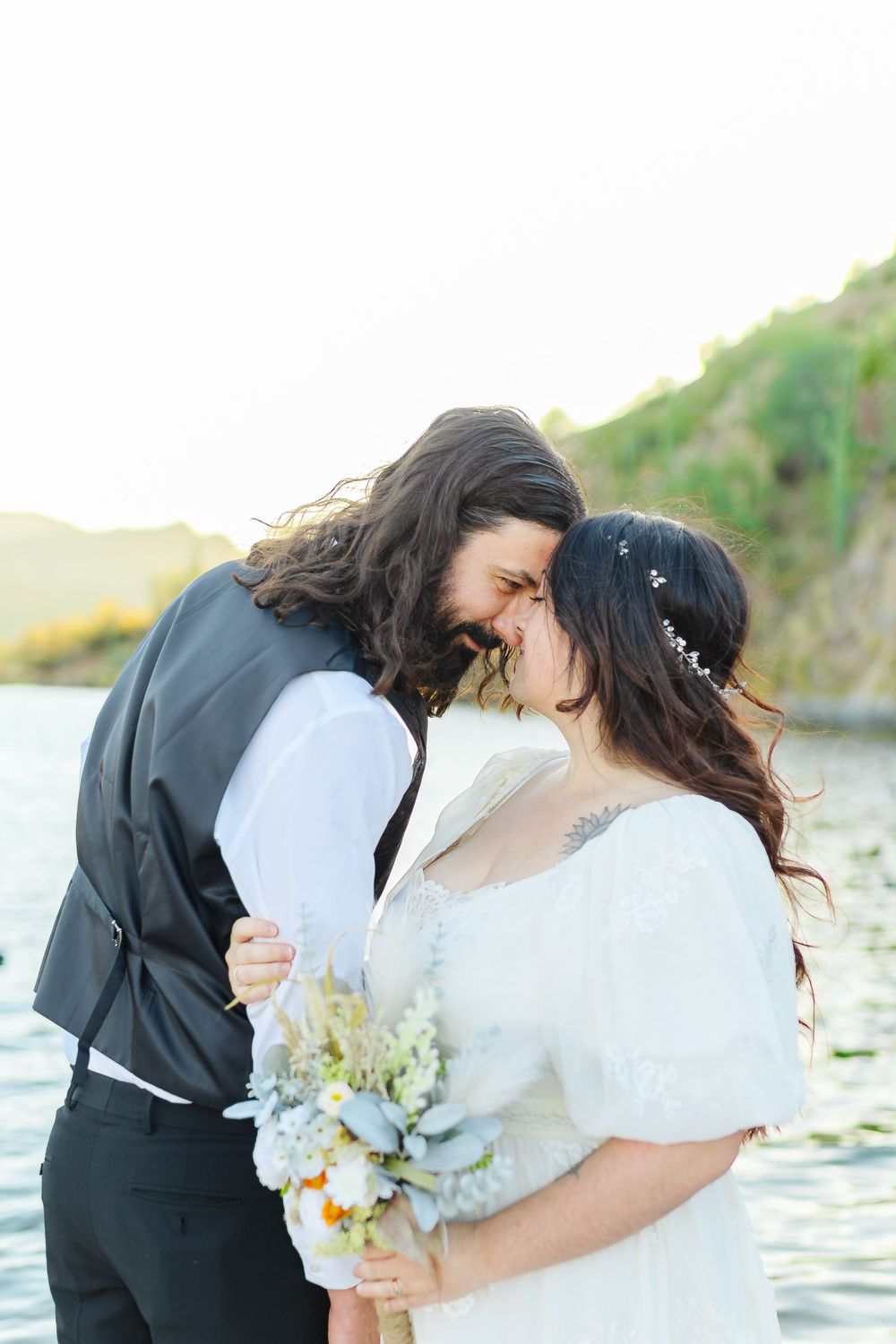 A newlywed couple touch foreheads. The wife holds a bouquet in one hand and holds her husbands arm with the other.