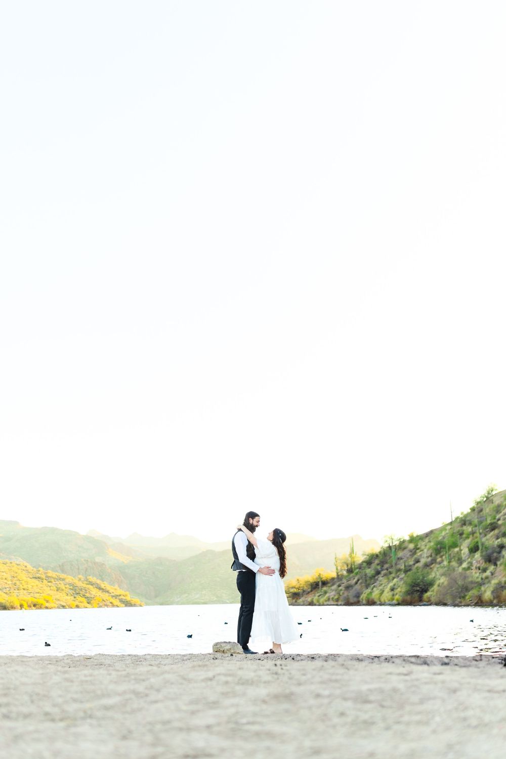 A newly married couple hold each other standing before a beautiful lake and mountain scenery.