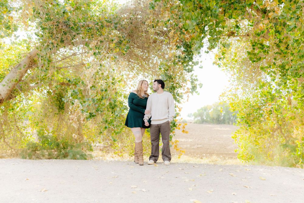 A young couple hold hands under green trees, looking into each other's eyes.