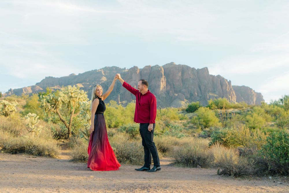 A couple stand before the Superstition Mountains. The wife is being twirled in a circle by her husband.