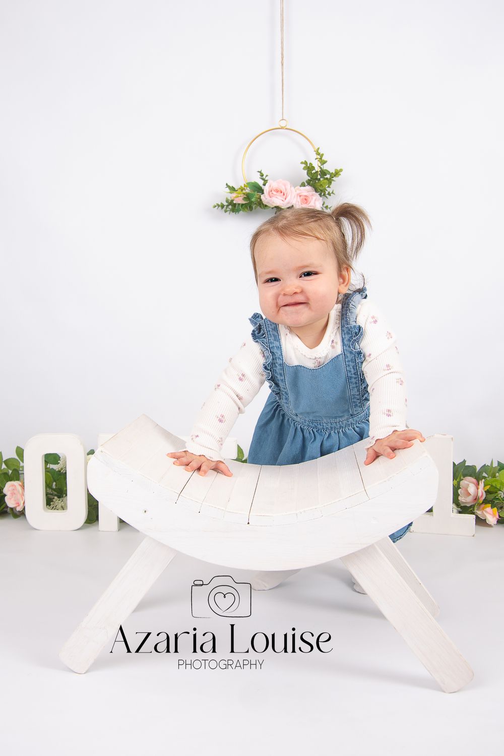 Small child wearing blue denim overalls and floral headband smiling while sitting on white wooden curved bench.