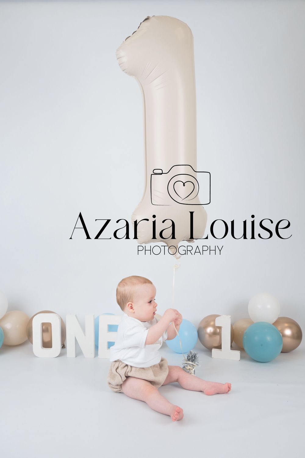 Studio photo of baby's first birthday setup with large number one balloon, blue and grey decorations against white backdrop.