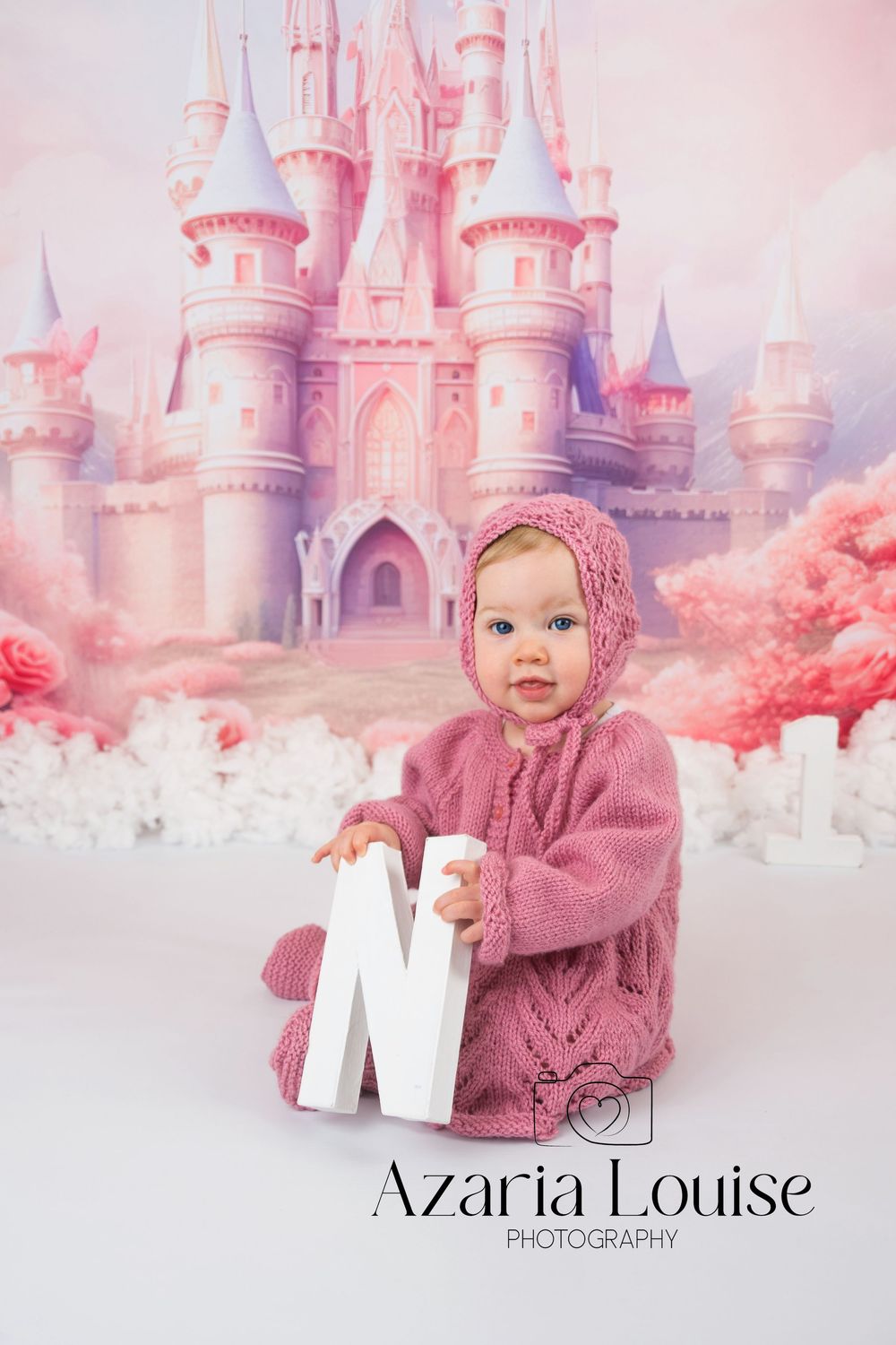 Child in pink knit sweater holds white letter N in front of dreamy fairytale castle backdrop with pink tones.