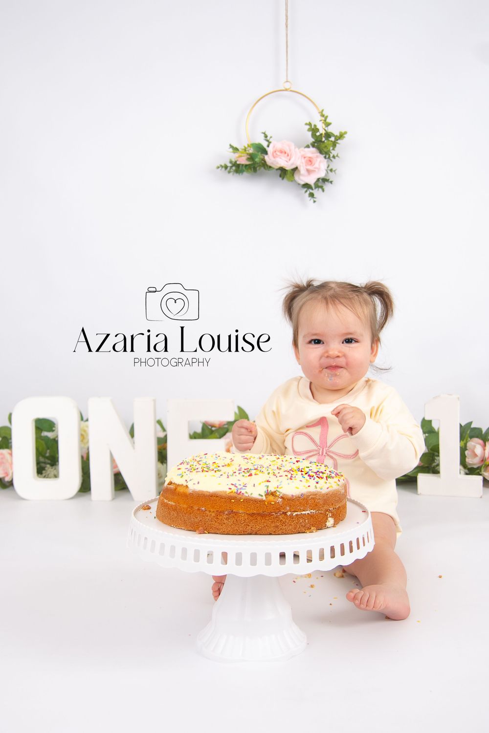 A baby in yellow sits at a cake smash photo session with white letters and floral decor on a white background.
