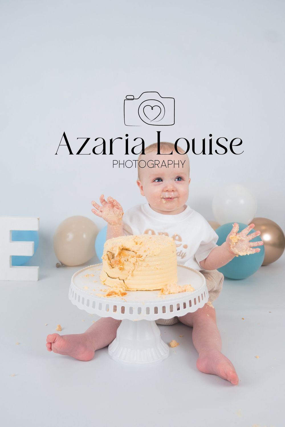 Baby in white outfit sitting with smashed orange birthday cake on pedestal stand, surrounded by blue and gray balloons.