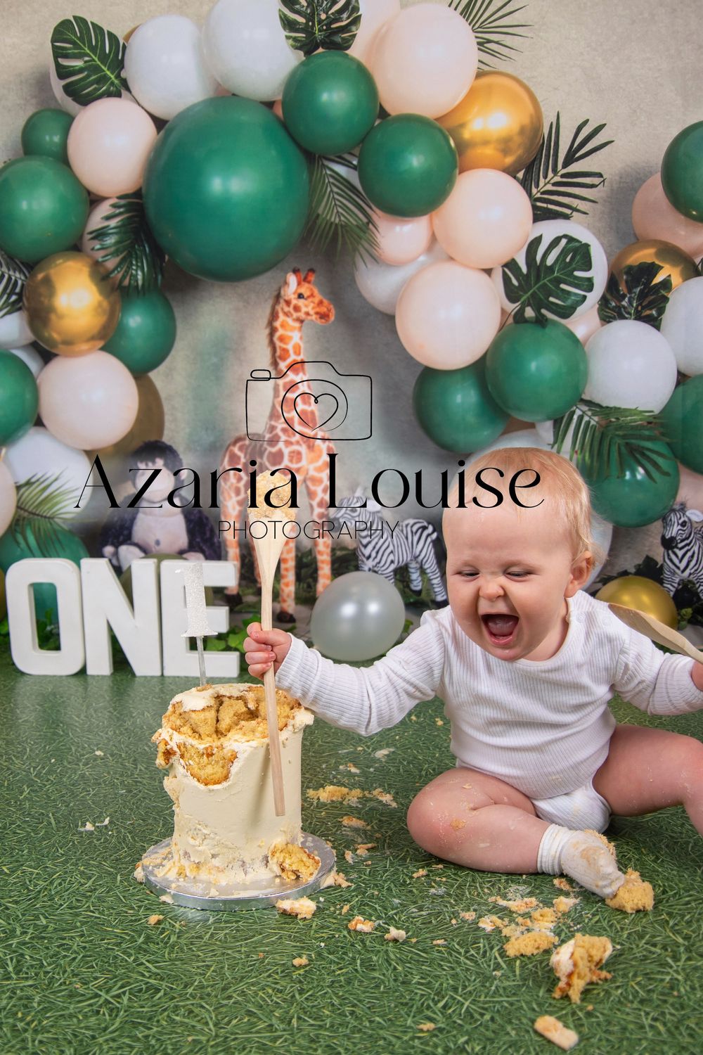 First birthday cake smash photo shoot with green and gold balloon arch, tropical leaves and a joyful moment captured.