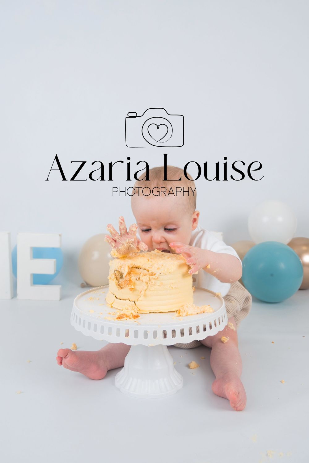 Baby sitting on white surface with cake smash setup featuring blue balloons and Azaria Louise Photography branding.