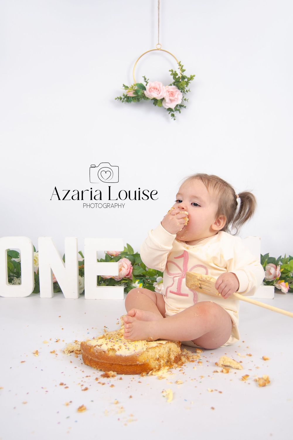 Baby in yellow outfit enjoying cake smash photo session with floral decorations and white ONE letters.