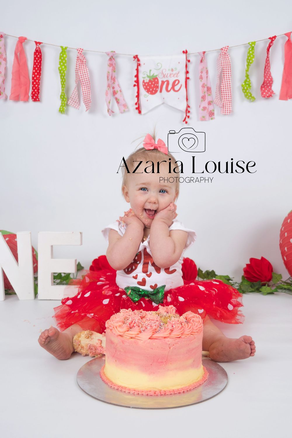 Pink and green fabric banner hangs above a small white chair and peachy pink ombre cake for first birthday photo session.