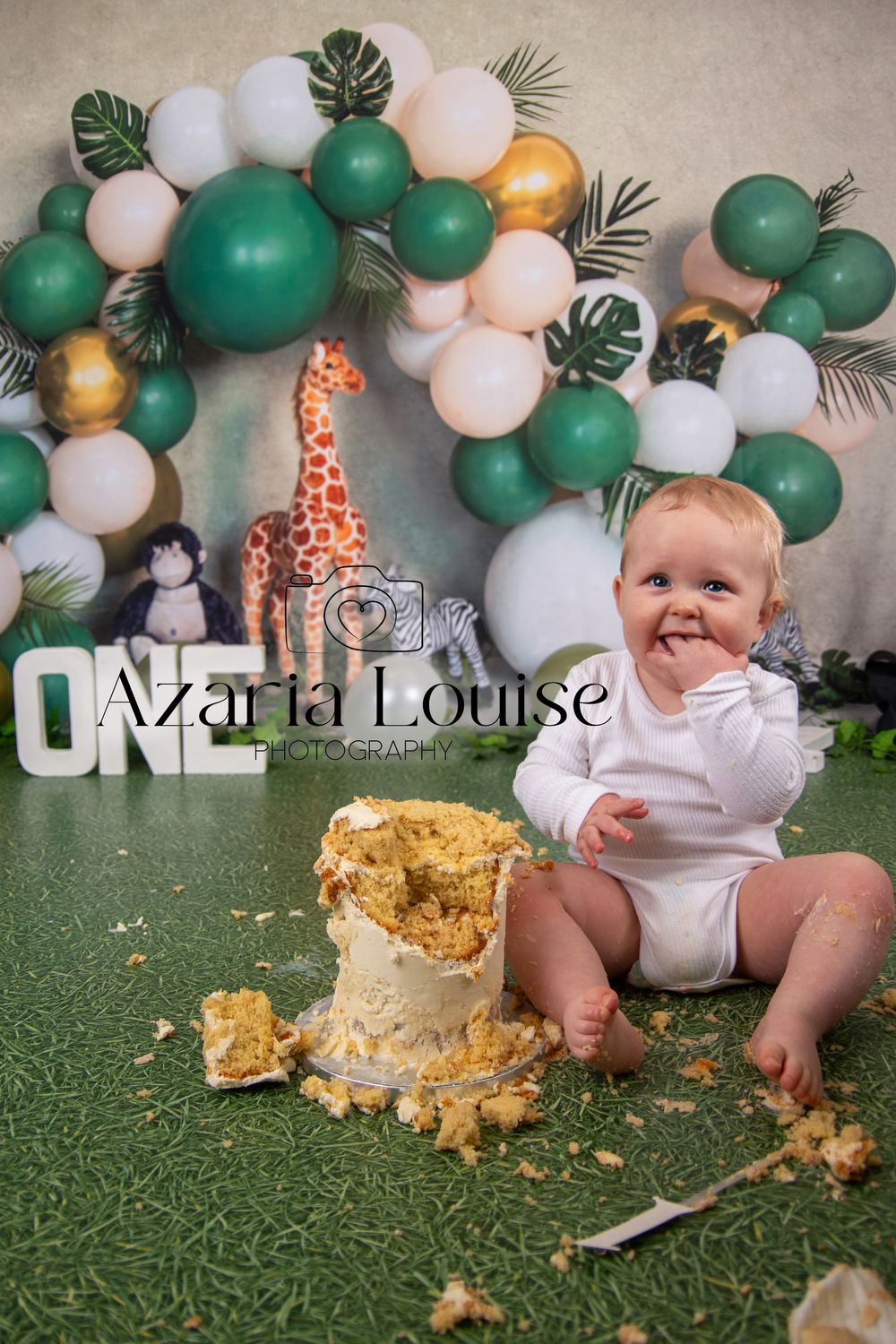 Jungle-themed first birthday party setup with green and white balloon arch, toy giraffe, and smashed cake on artificial grass.