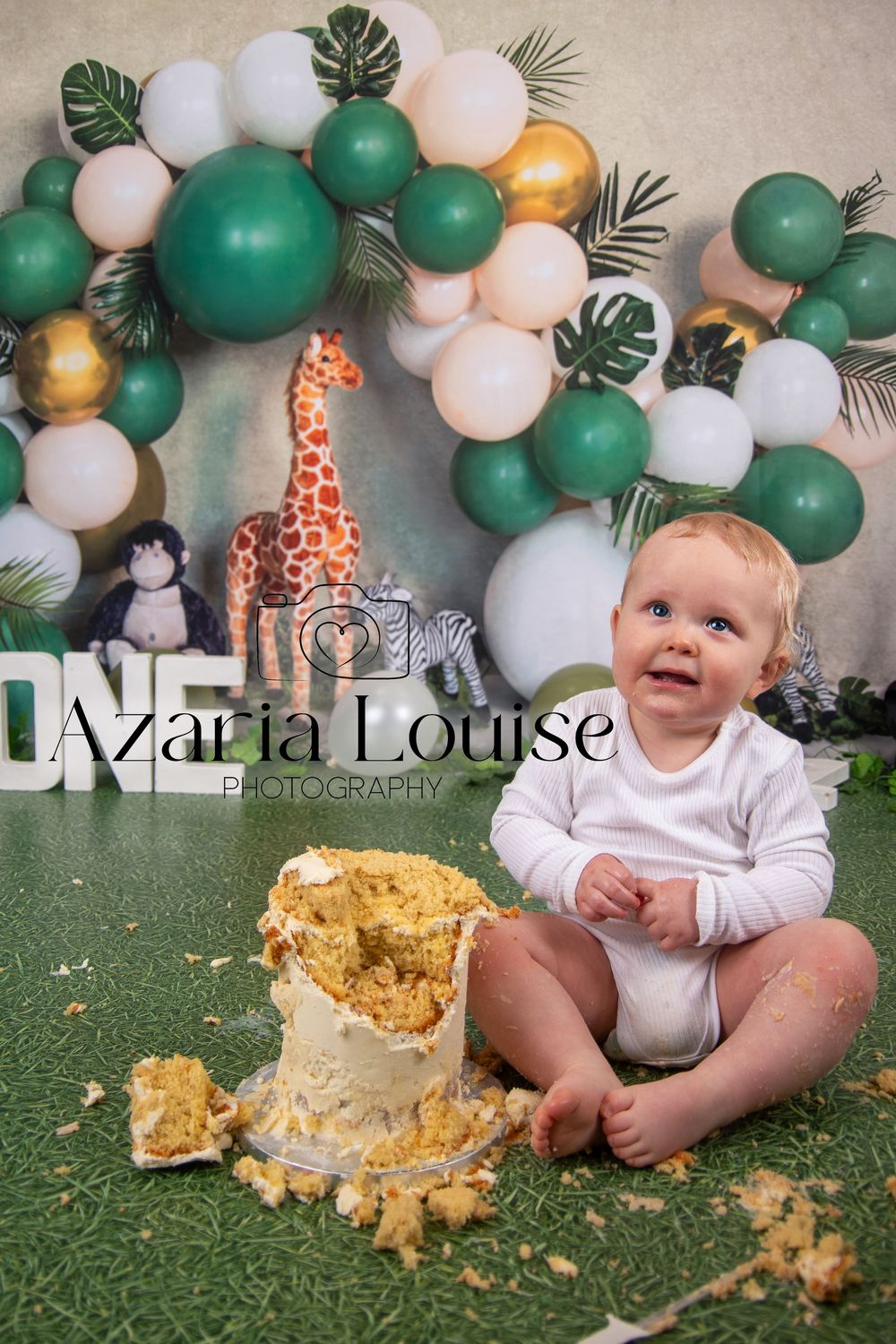 First birthday cake smash photo with safari theme featuring green and white balloon arch and toy giraffe decoration.