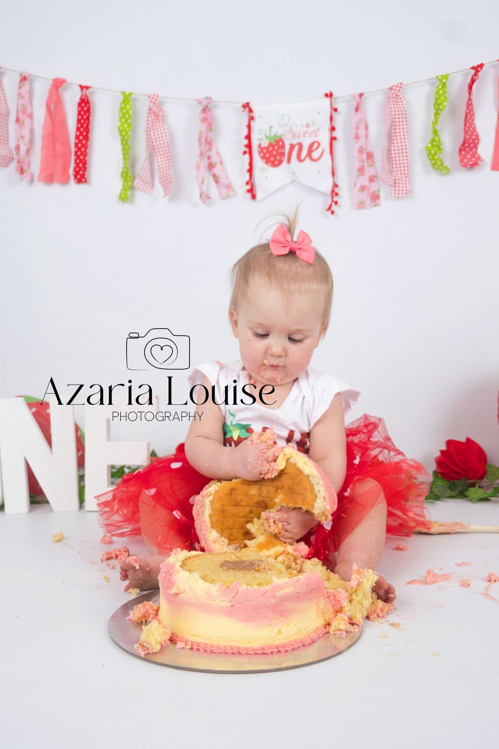 Pink and coral cake smash photography session with fabric banner and tutu decorations against white background.