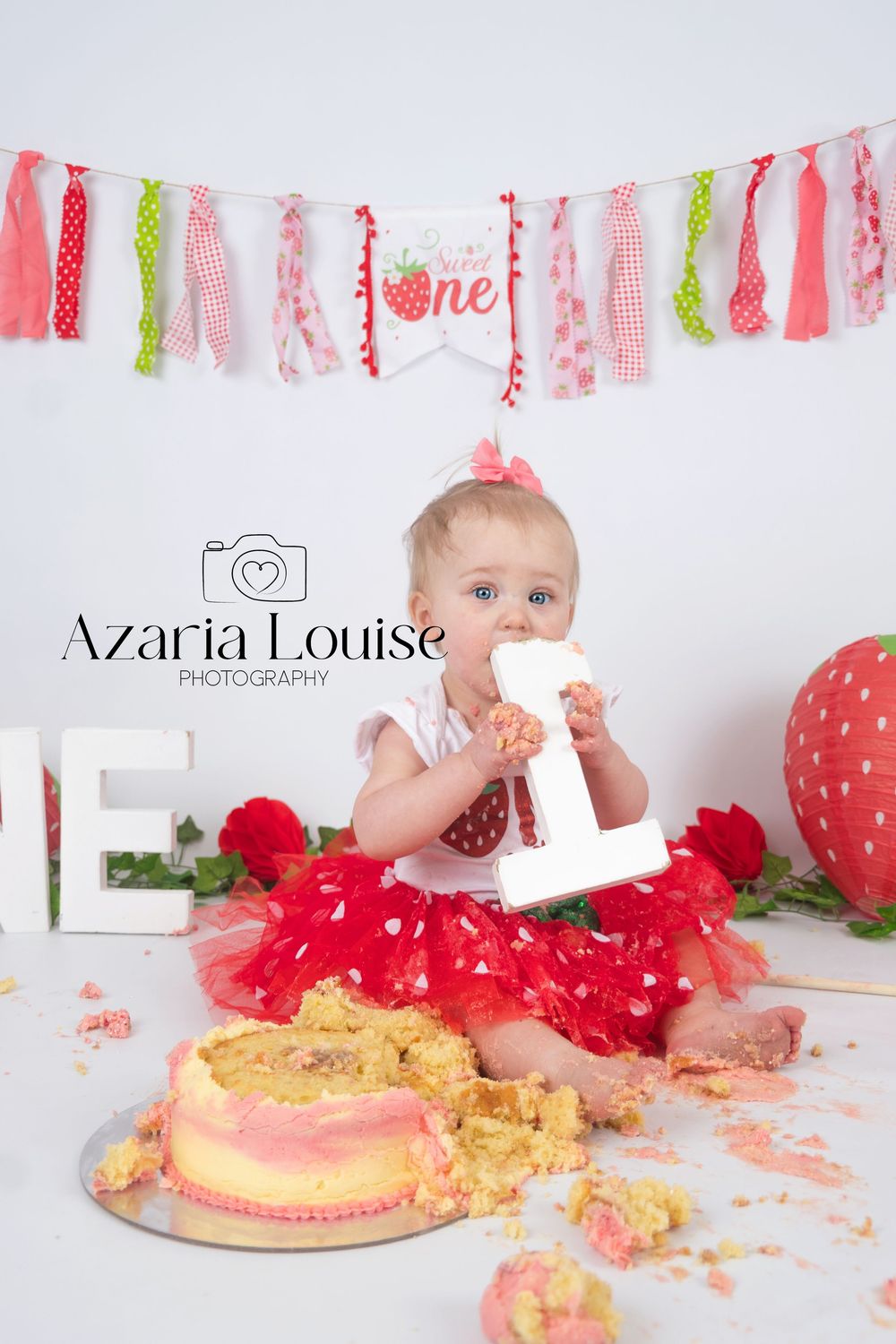First birthday cake smash photo shoot with pink and green banner, red tutu, and strawberry themed decorations.