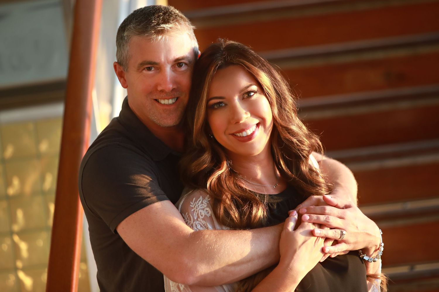 A couple embraces and smiles warmly while posing together against an orange wall background.