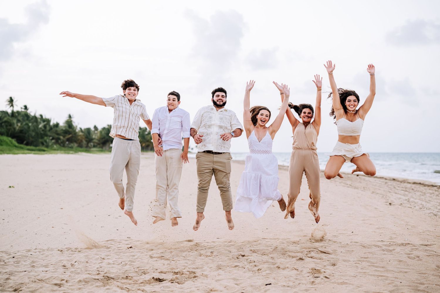 Wedding party jumping joyfully on beach in coordinated beige and white outfits.
