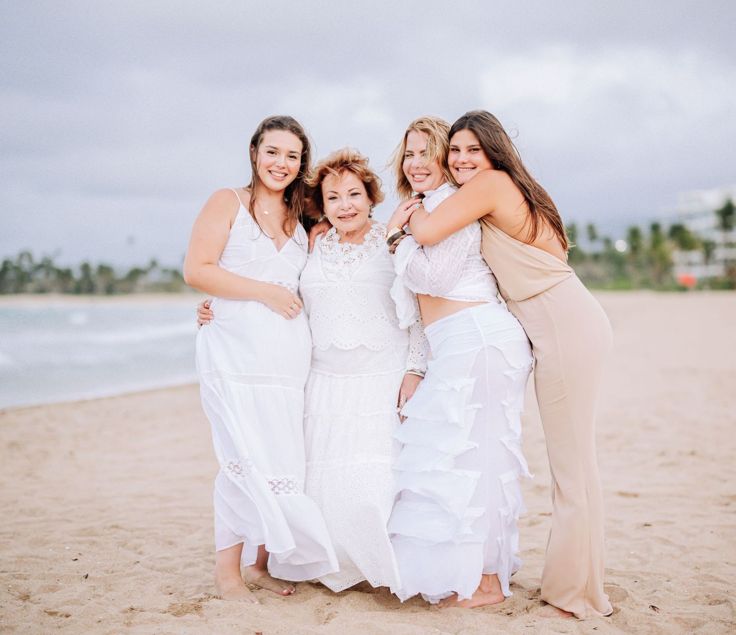 Group hugging and smiling together on sandy beach in white dresses.
