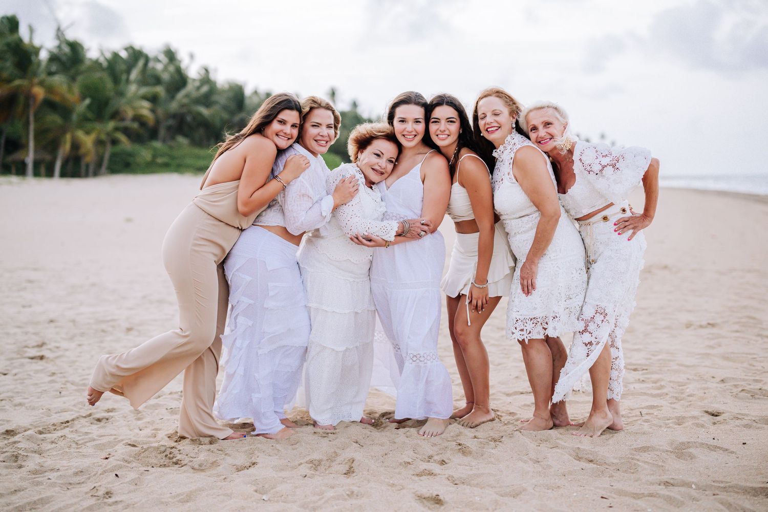 Friends embracing and laughing together on tropical beach in white and beige outfits.